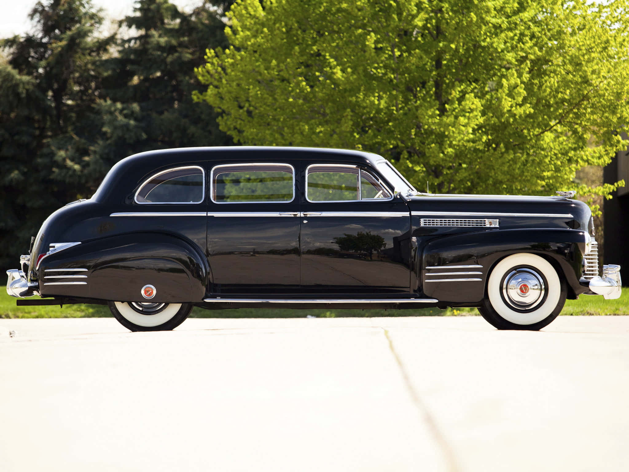 Cadillac Fleetwood 桌布 壁紙 — 一輛光澤黑色復古轎車配白色輪胎，在明媚陽光下與茂密綠樹旁閃閃發光。