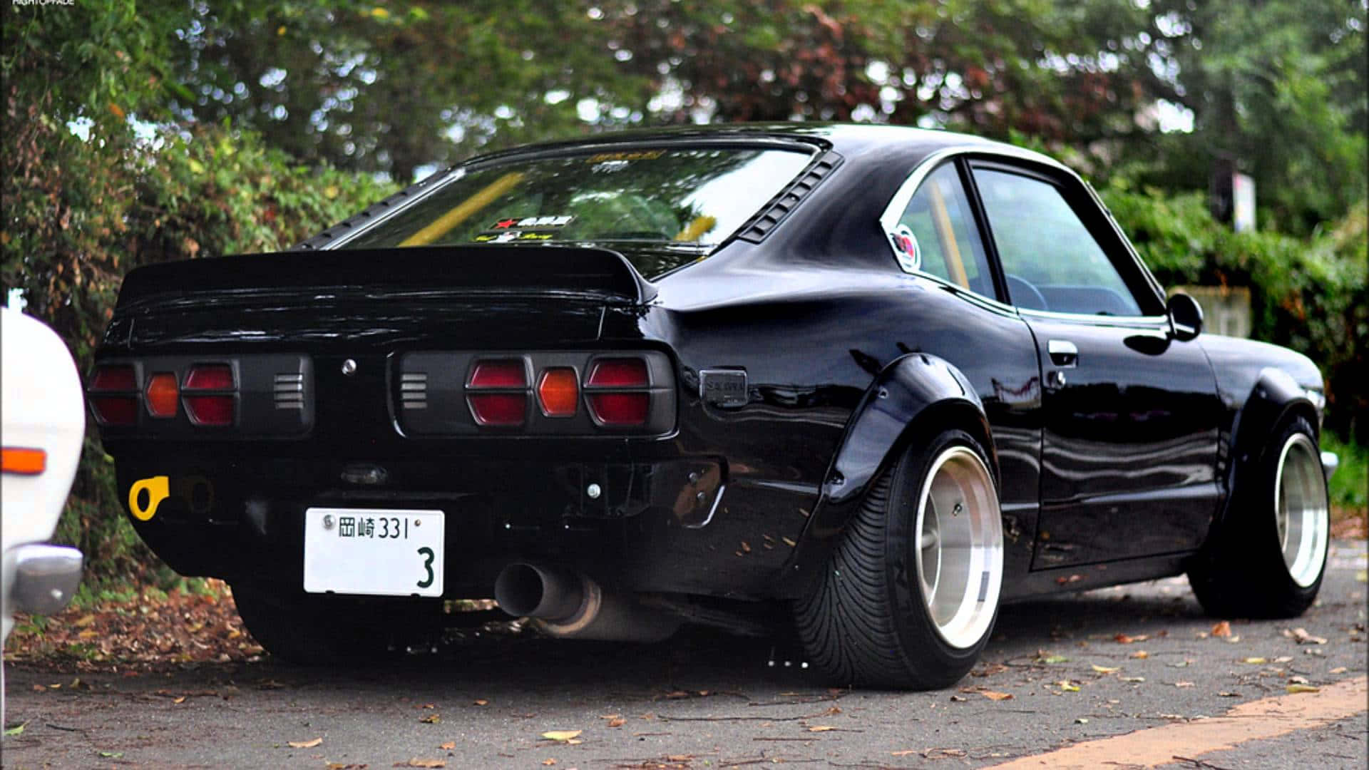 Mazda Rx-3 桌布 壁紙 — 一輛光澤黑色經典日本跑車停在綠葉成蔭的路邊，配備寬大的白色輪圈和紅色尾燈。