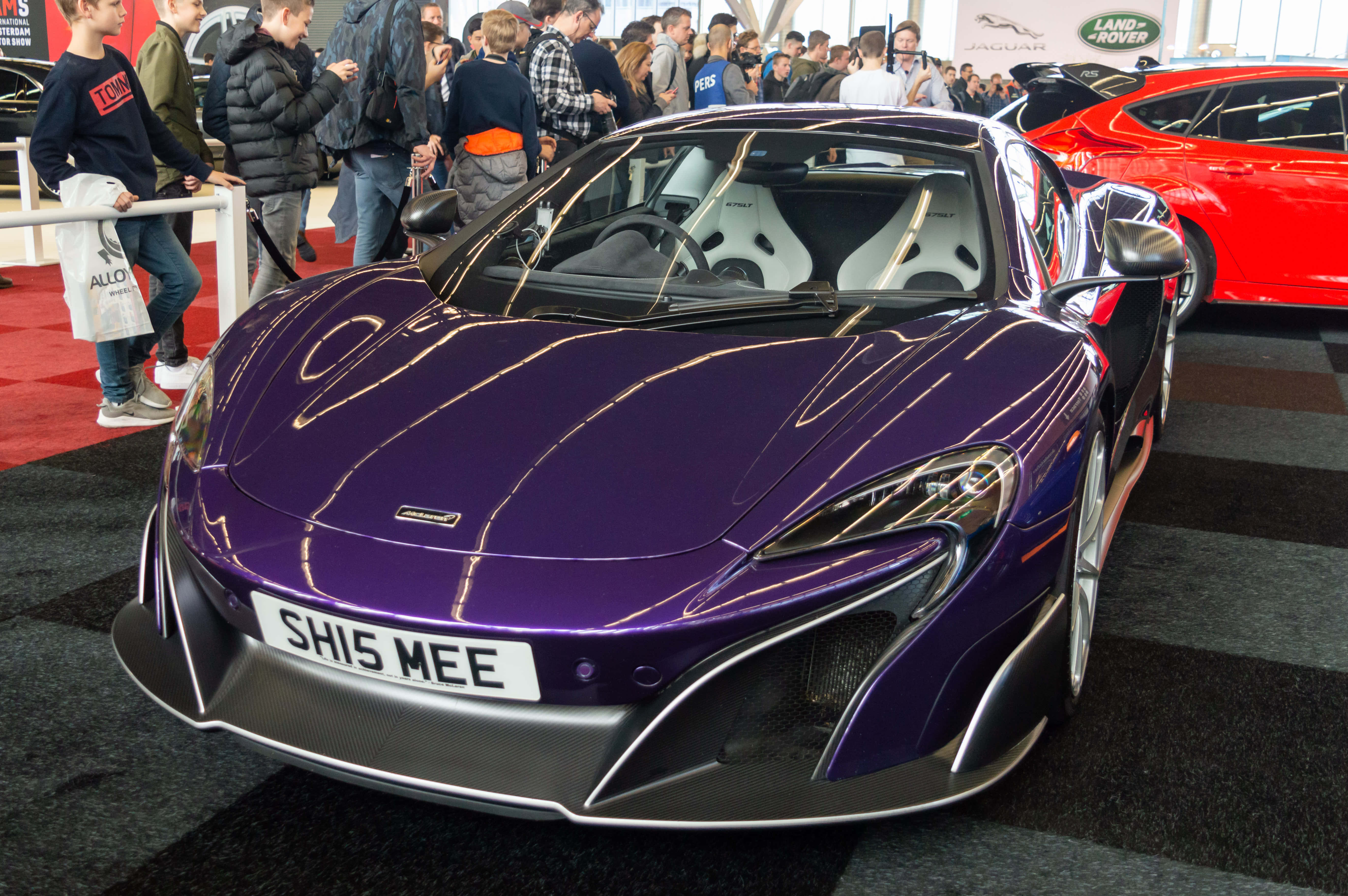 Mclaren 675lt Spider 桌布 壁紙 — 一輛光澤的紫色麥拉倫跑車在擁擠的車展上，在明亮的燈光下閃閃發光，觀眾欣賞其時尚設計。