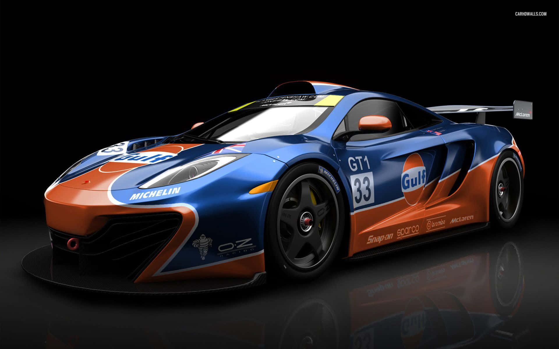 Mclaren F1 桌布 壁紙 — 一輛光滑的藍色和橙色海灣塗裝麥拿倫賽車，編號33，在攝影棚燈光下於反光黑色表面上閃閃發光。