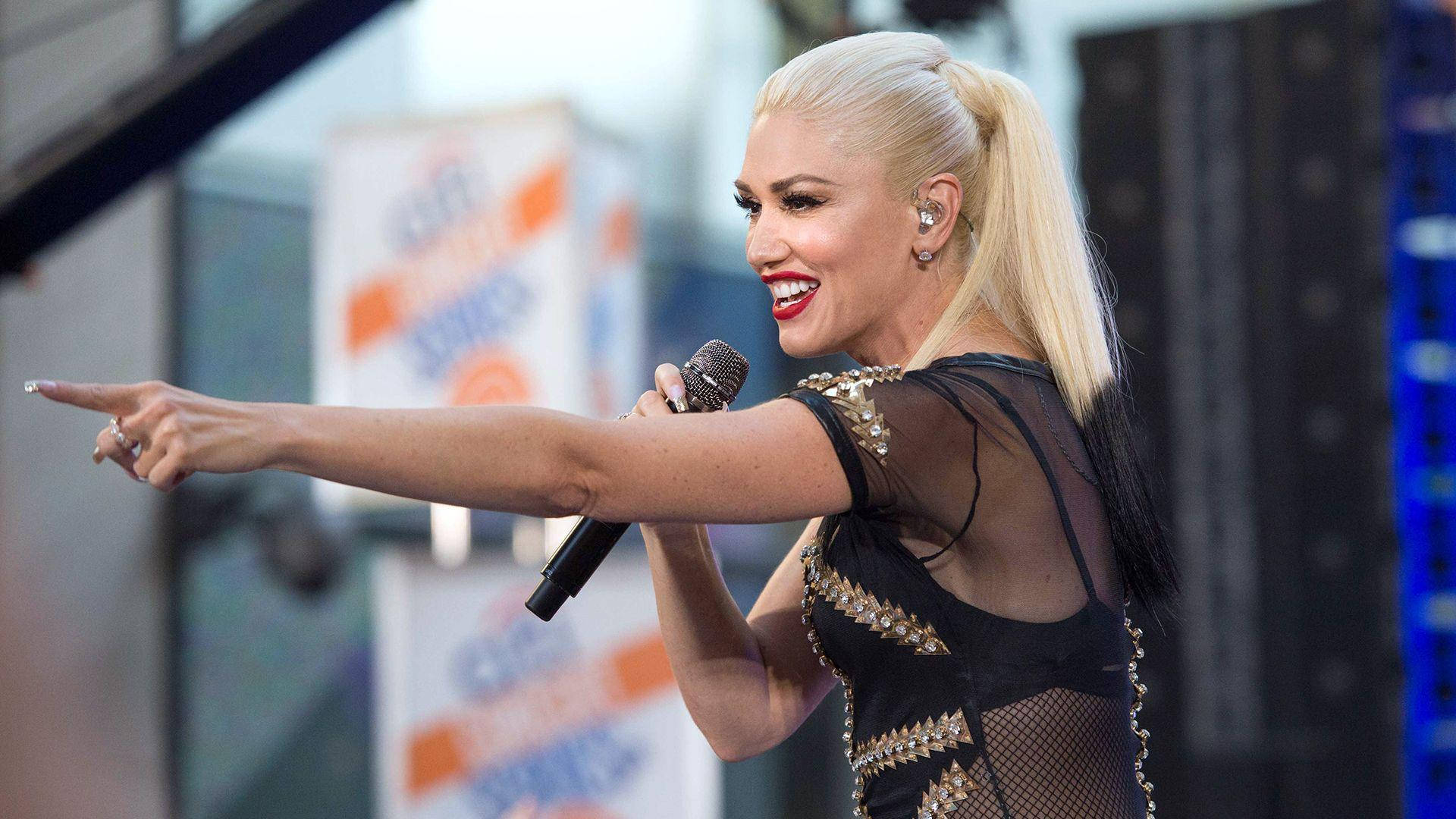 Stefani 桌布 壁紙 — Gwen Stefani 在舞台上穿著黑色鉚釘服裝，充滿活力地指向人群，露出燦爛的笑容。