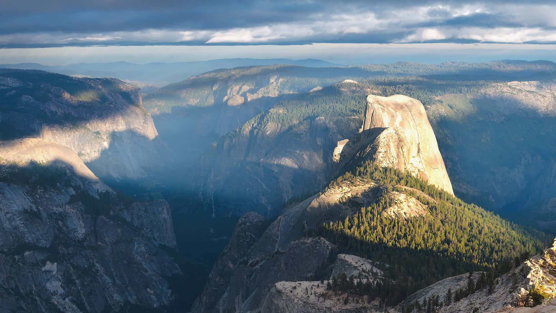 1080p 桌布 壁紙 — Half Dome 聳立於霧氣瀰漫的山谷之上，陽光照射的花崗岩懸崖和茂密的松林在戲劇性的雲層下。