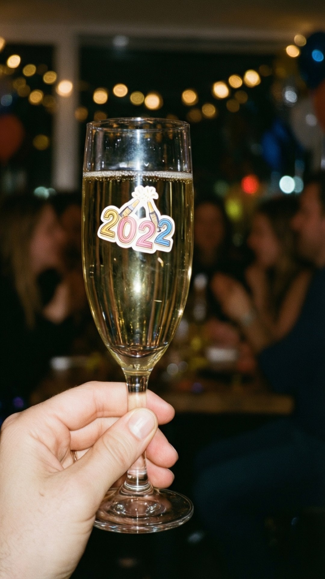 Champagne Toast Sticker 2022 壁紙 — 一隻手拿著一個帶有2022年除夕貼紙的香檳杯，背景是模糊的派對場景和散景燈光。