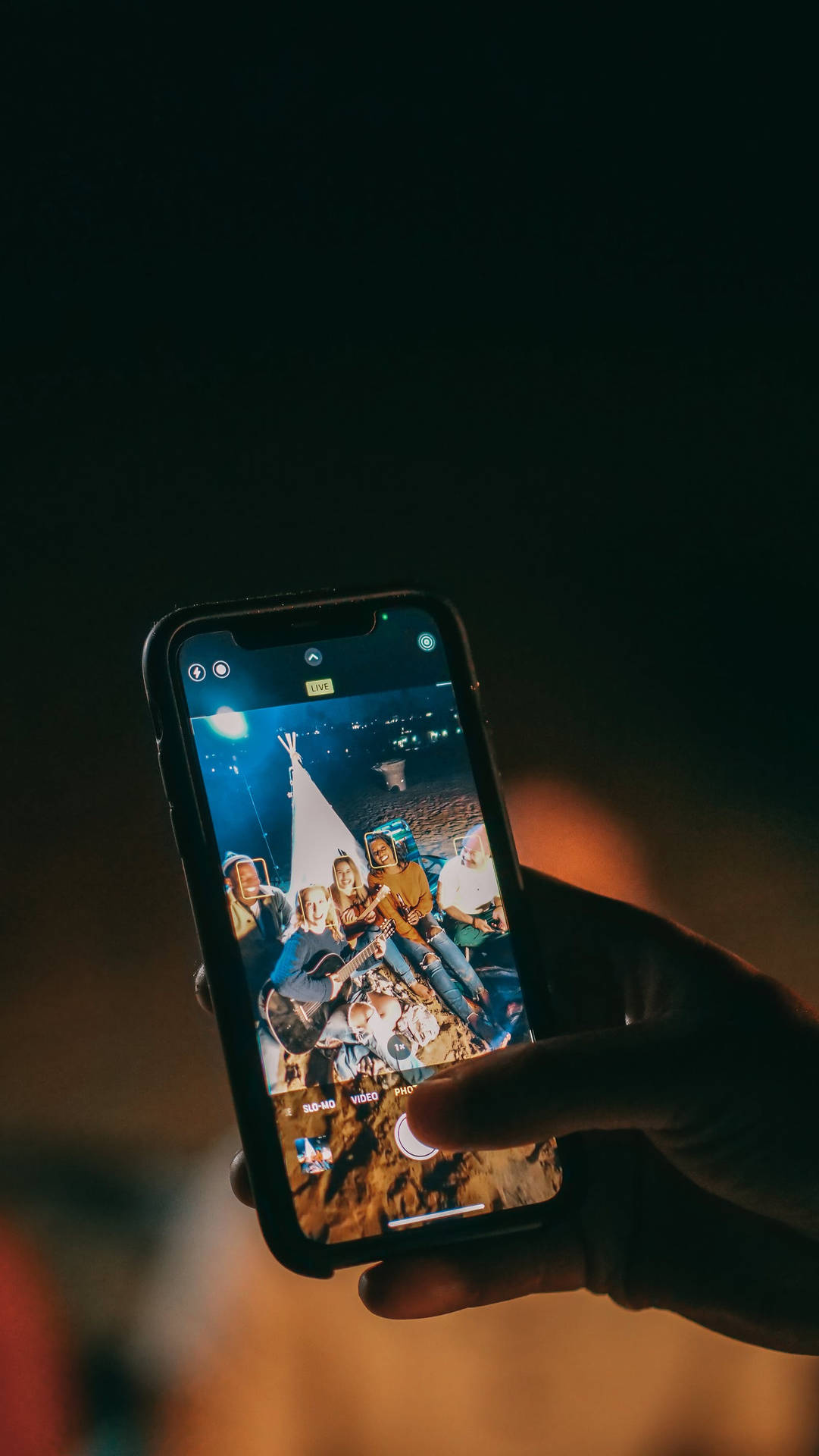手拿著手機，顯示朋友們在夜晚圍著火露營。 — iPhone 11 壁紙