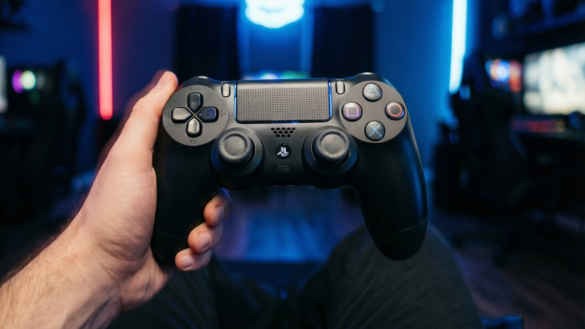 Holding PS4 Controller 壁紙 — 一隻手在昏暗的房間中握著黑色PlayStation DualShock控制器，房間被藍色和紅色的霓虹光籠罩。