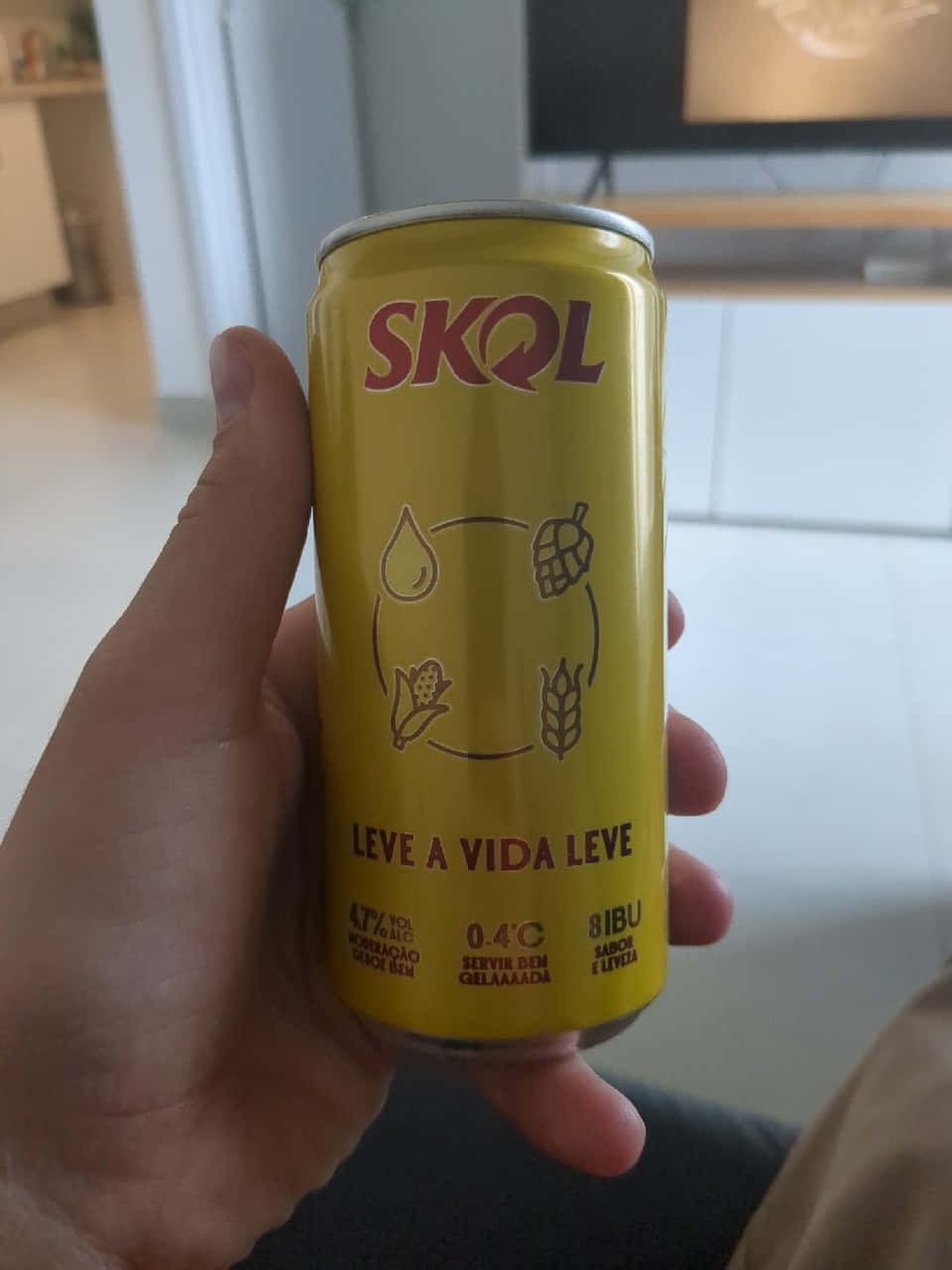  壁紙 — 一隻手拿著一個亮黃色的Skol啤酒罐，上面有啤酒花和小麥圖標，背景是模糊的室內環境。
