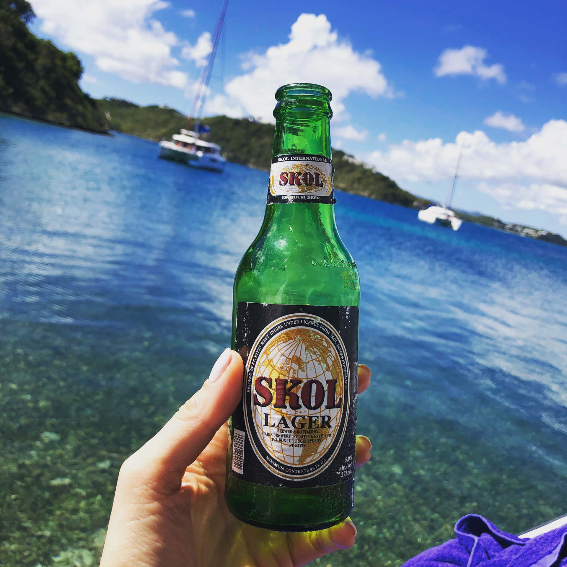  壁紙 — 一隻手拿著一瓶冰涼的綠色Skol Lager啤酒，背景是碧藍的海水、帆船和翠綠的山丘，在明亮的藍天下。