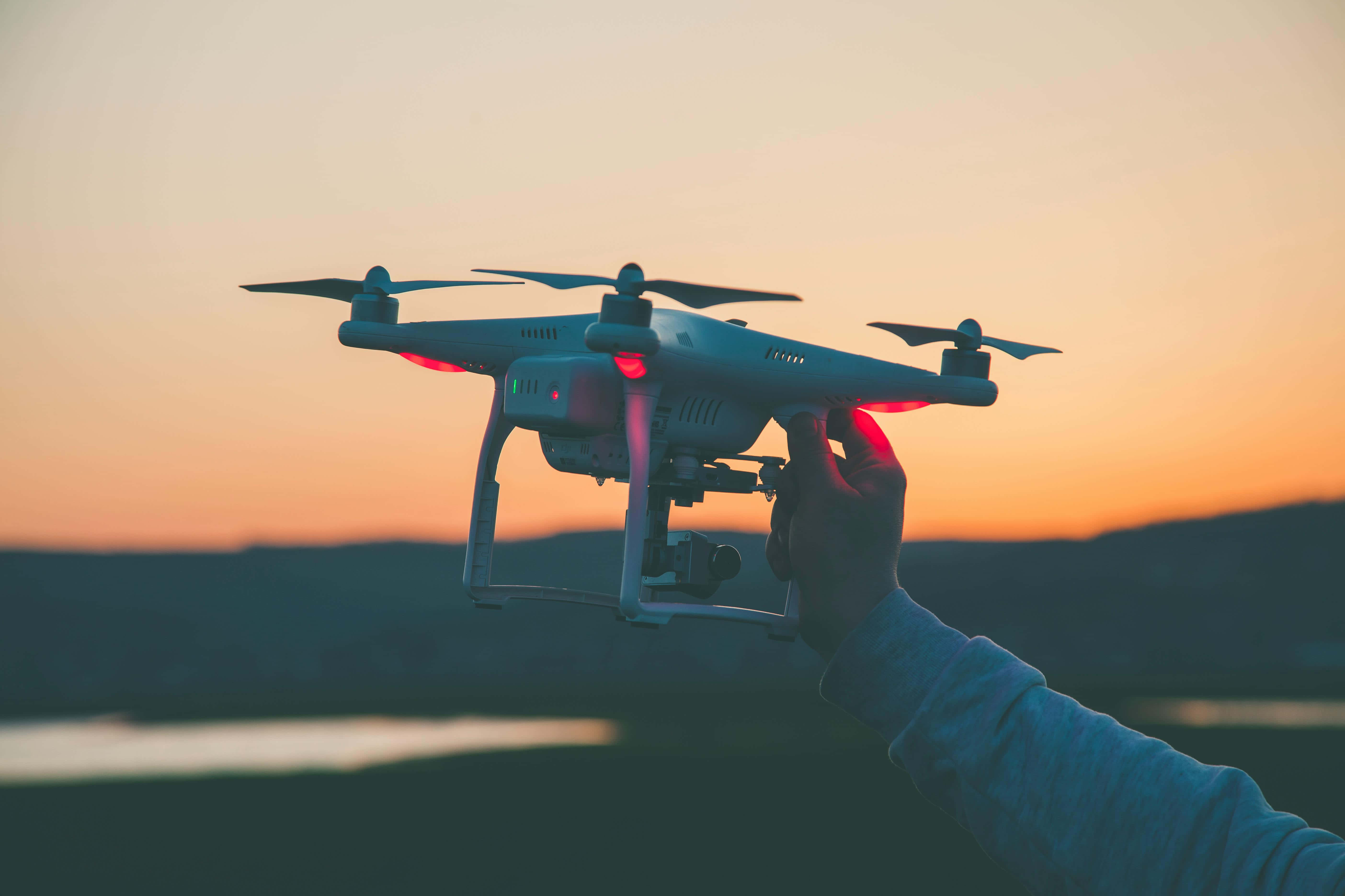 Drone Camera 桌布 壁紙 — 一隻手在溫暖的日落天空下拿著無人機，有發光的紅燈和遠處的山丘。