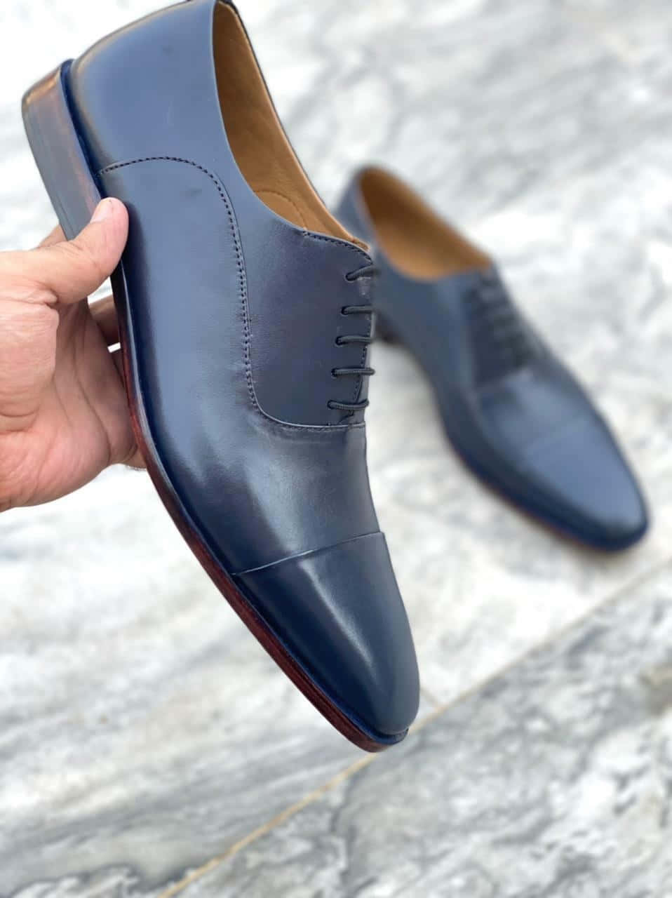 Navy Blue Scarf 桌布 壁紙 — 一隻手拿著一雙時尚的深藍色 cap-toe 牛津鞋，搭配酒紅色鞋底，背景為大理石地板。