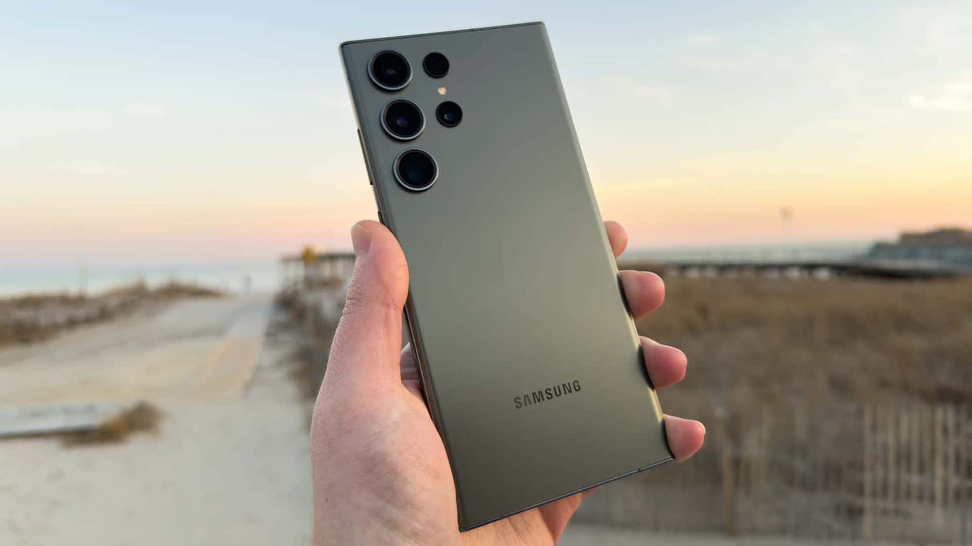  壁紙 — 一隻手拿著一款時尚的橄欖綠色Samsung智慧型手機，背景是柔和的海灘日落，帶有模糊的沙丘和海洋地平線。