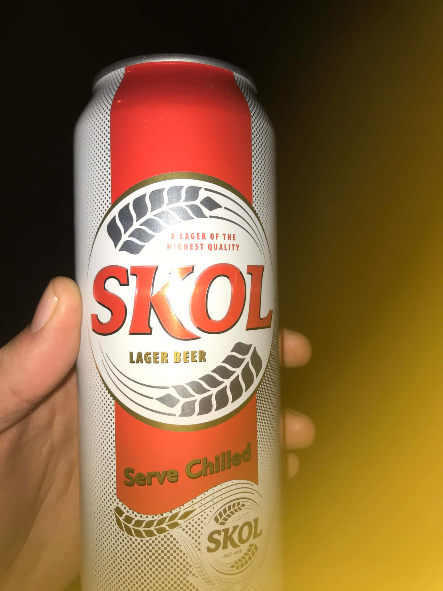  壁紙 — 一隻手拿著一個紅銀色的Skol拉格啤酒罐，背景是溫暖的黃色漸變。