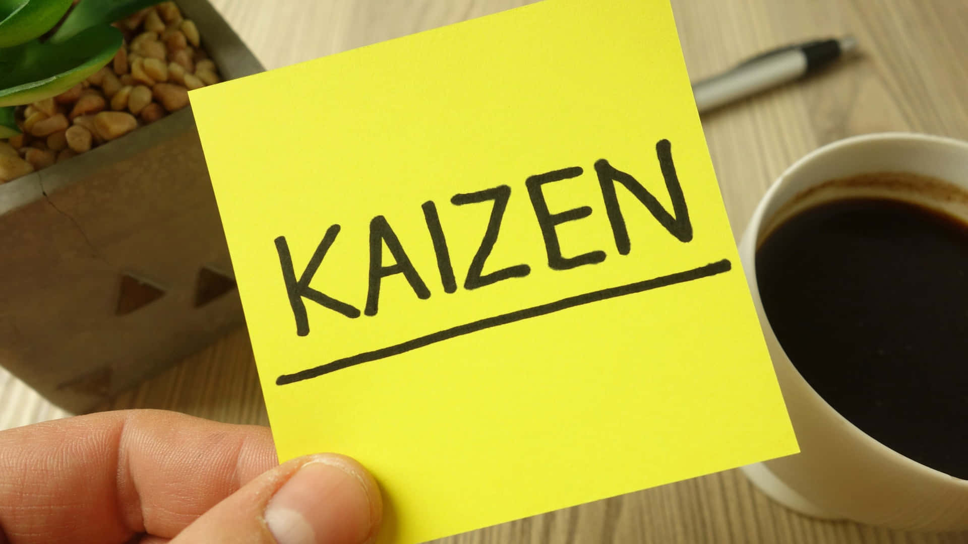  壁紙 — 一隻手拿著一張黃色便利貼，上面用粗黑馬克筆寫著「KAIZEN」，旁邊有一杯咖啡和一盆多肉植物放在木桌上。