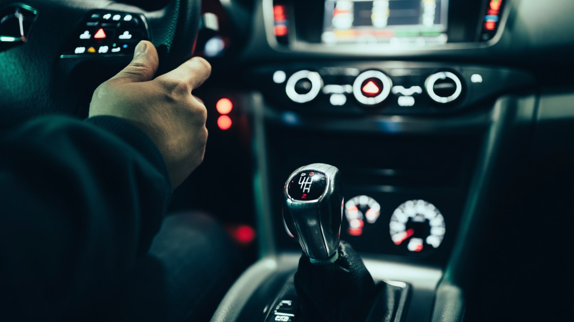 Gear Shift Focus 壁紙 — 一隻手抓住方向盤，在黑暗的車內，聚焦於發光的紅色和白色儀表板燈光以及手動變速桿。