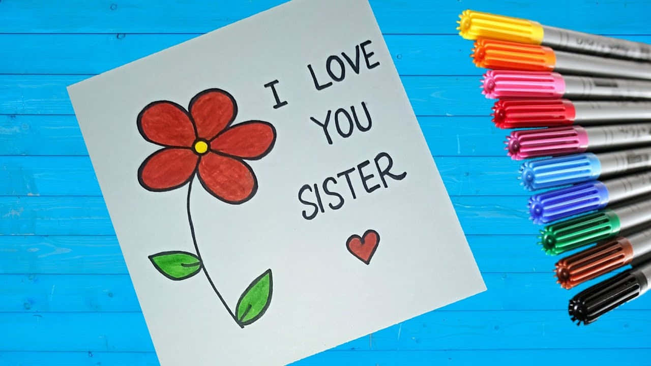  壁紙 — 一張手工卡片，上面有紅色花朵和「I LOVE YOU SISTER」文字，放在藍色木質表面上，旁邊有彩色記號筆。