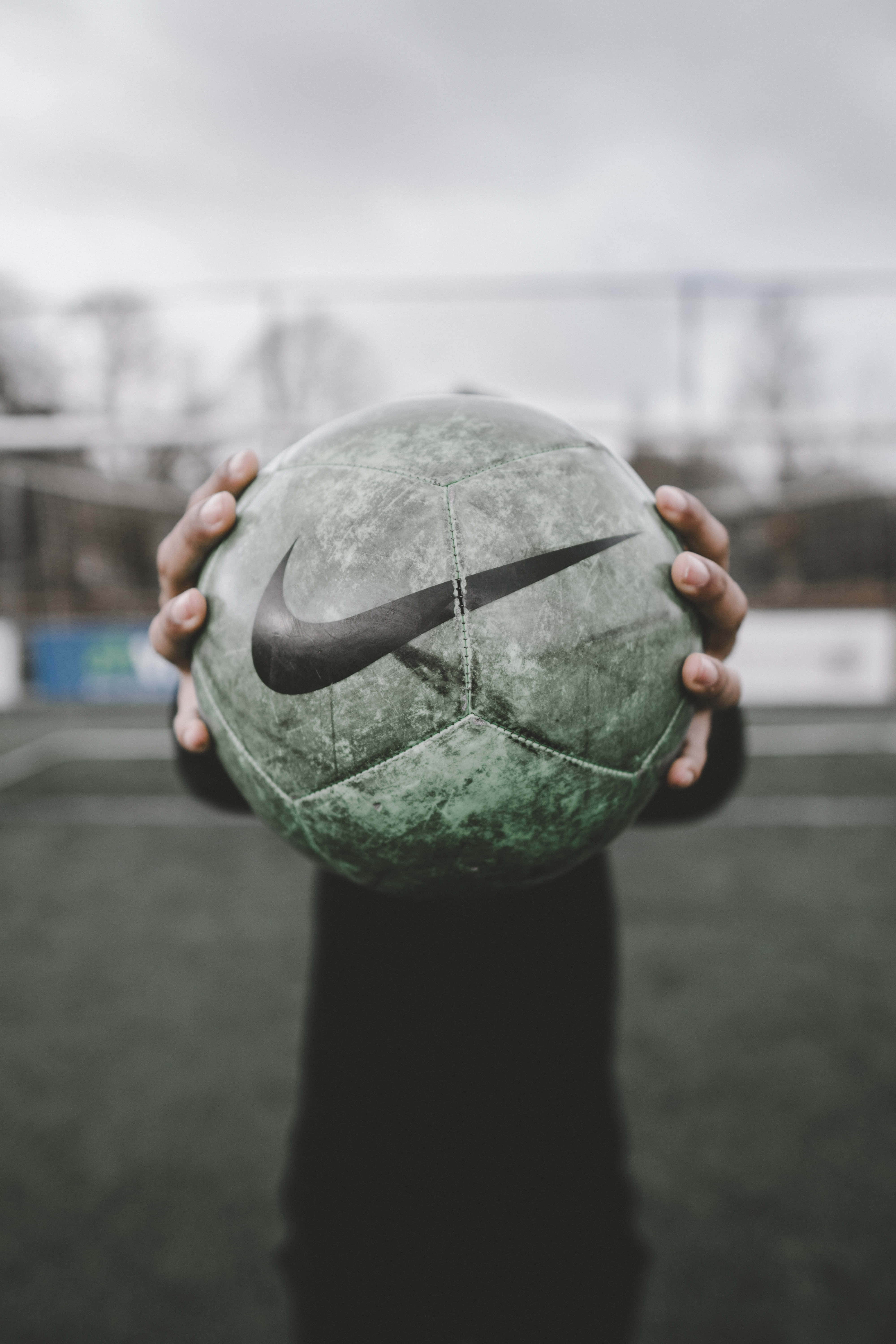 ball 桌布 壁紙 — 雙手在陰天的戶外球場上，拿著一個風化了的綠色Nike足球，背景模糊。