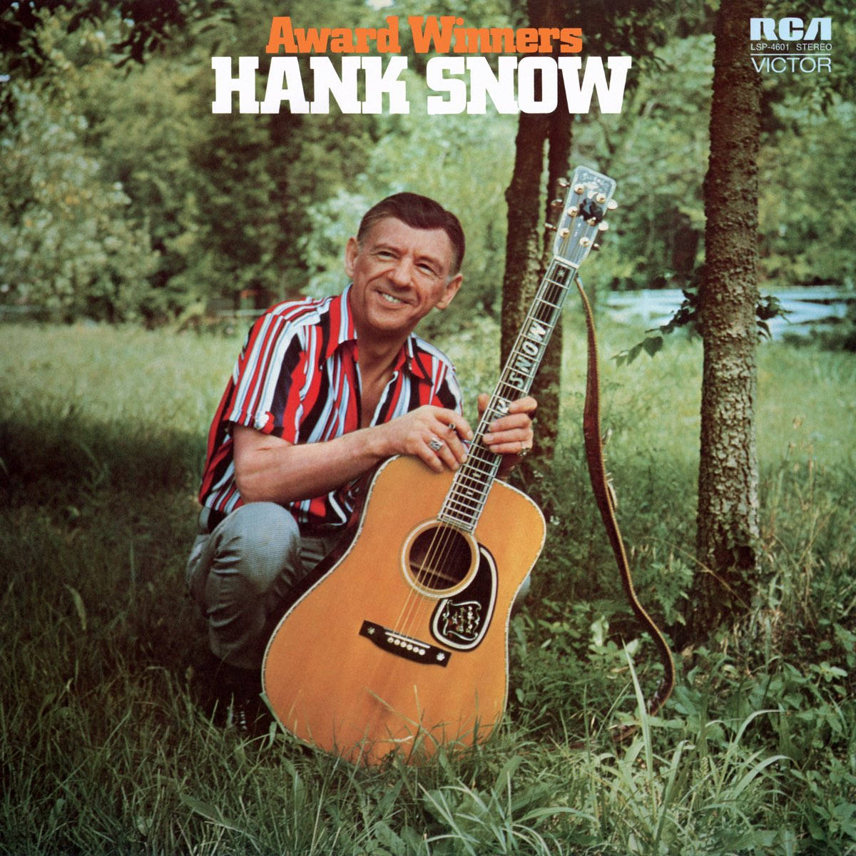 音樂 桌布 壁紙 — Hank Snow 在陽光明媚的戶外環境中，跪在草地上，手持木吉他，在樹旁露出溫暖的笑容。