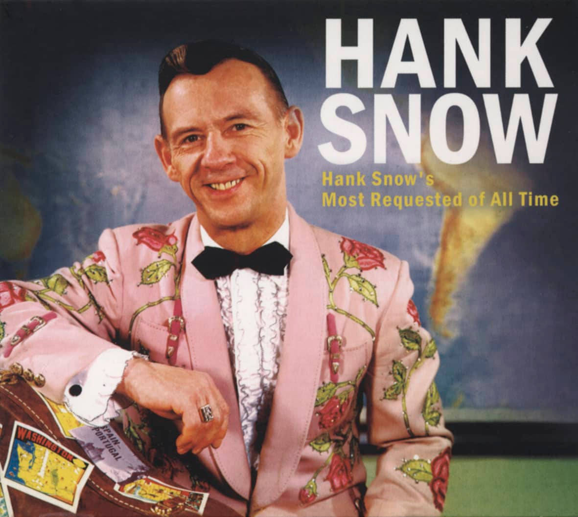 音樂 桌布 壁紙 — Hank Snow 身穿粉紅色玫瑰刺繡西裝，微笑著拿著一個貼有旅行貼紙的手提箱。