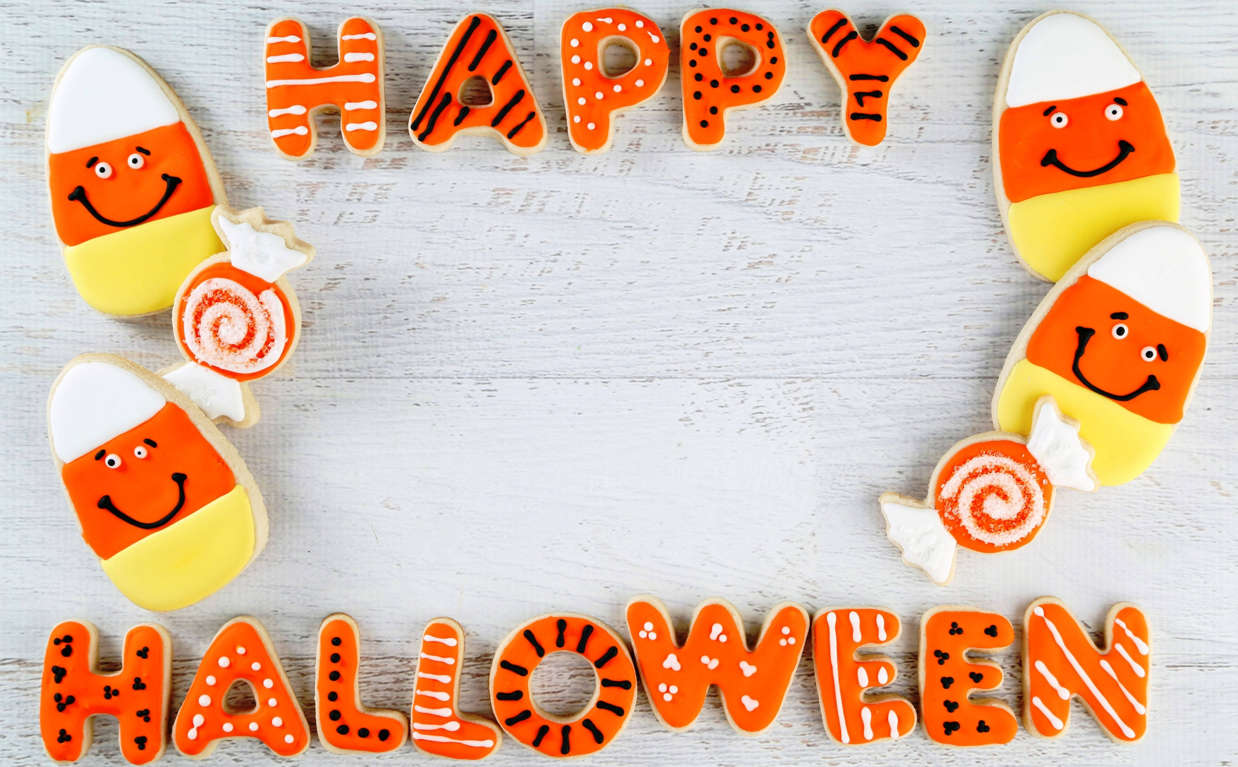 可愛萬聖節 桌布 壁紙 — Happy Halloween 用橙色裝飾餅乾拼出，餅乾有糖果玉米和棒棒糖形狀，放在白色木質表面上。