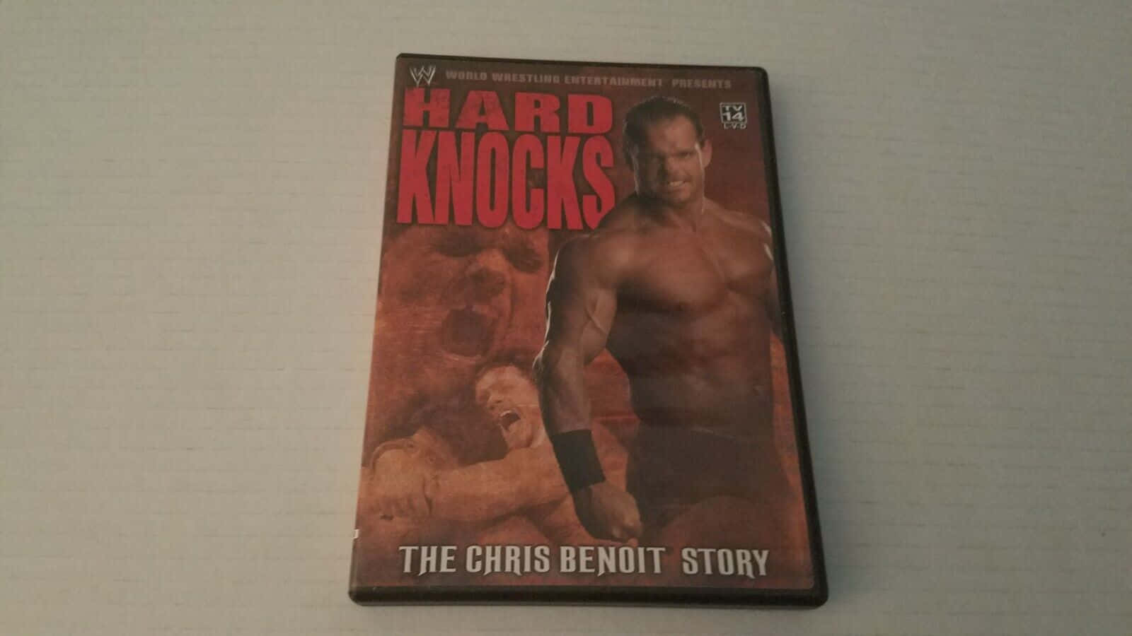 Story 桌布 壁紙 — 《Hard Knocks: The Chris Benoit Story》的DVD封面，展示一位肌肉發達的摔角手站在倒下的對手之上，背景是火紅色的。