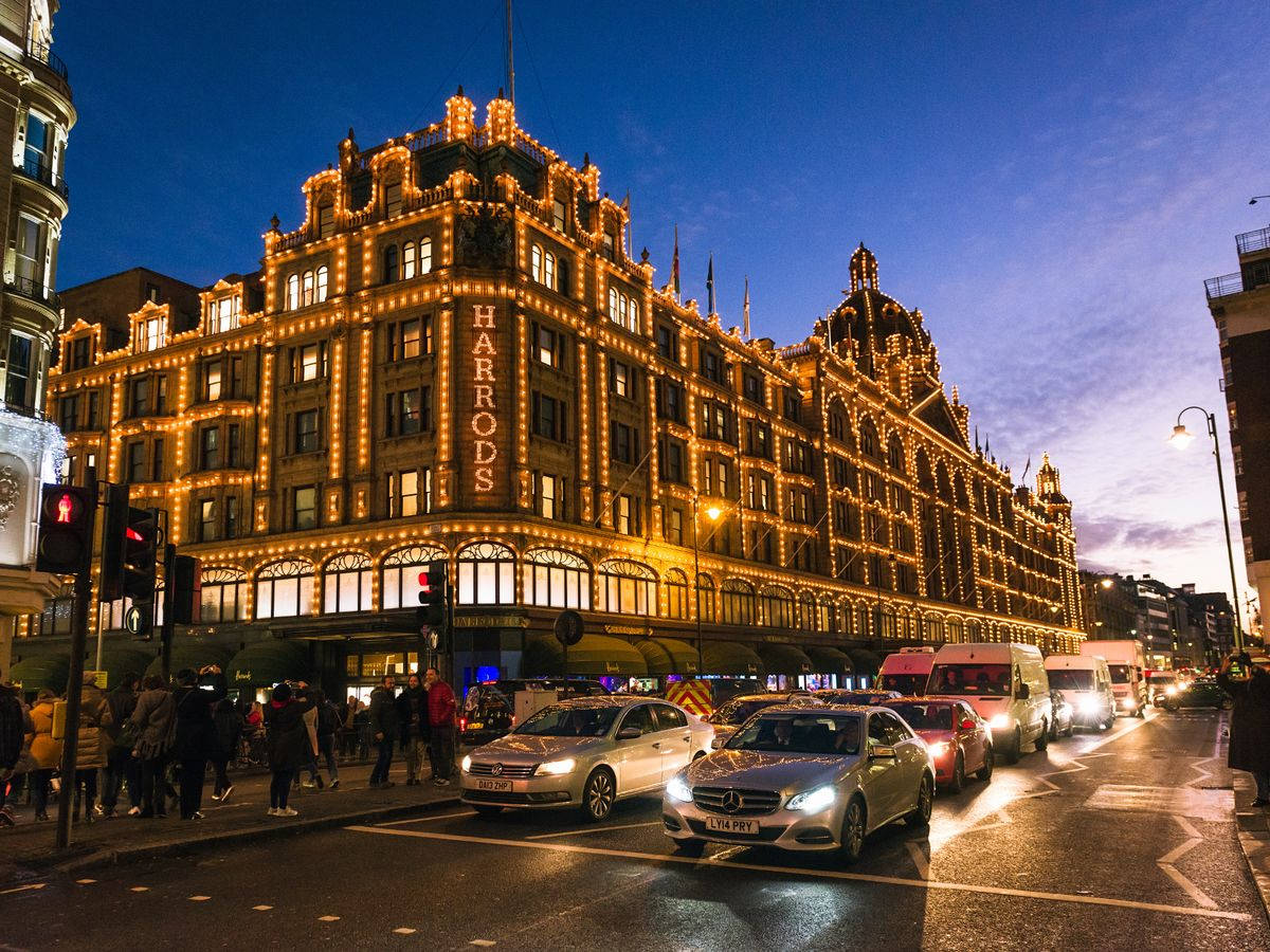 倫敦 桌布 壁紙 — Harrods 百貨公司在暮色天空下溫暖地發光，汽車和行人沿著潮濕的城市街道移動。