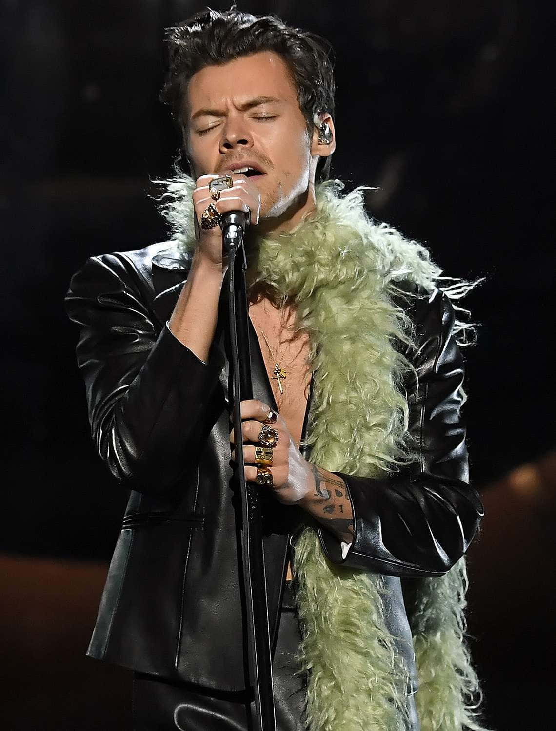 Harry Styles 2021 桌布 壁紙 — Harry Styles 熱情地對著麥克風唱歌，身穿黑色皮夾克和綠色毛皮披肩，在舞台燈光下。