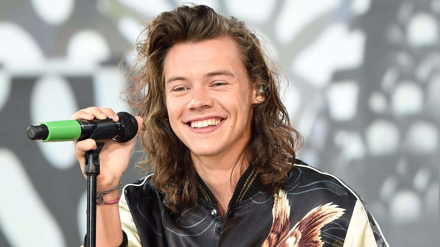 哈利·史泰爾斯 桌布 壁紙 — Harry Styles 燦爛地笑著，在舞台上拿著綠色麥克風，背景是模糊的抽象圖案。