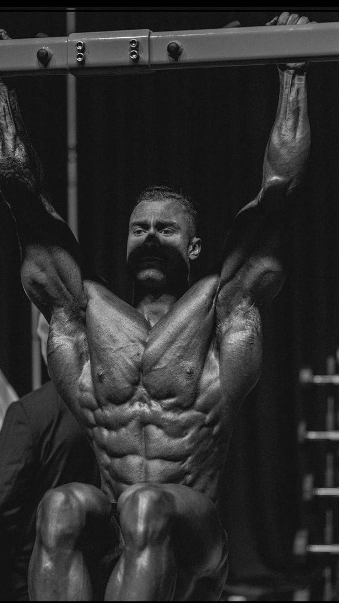 一名留鬍鬚的男子在健身房做引體向上時繃緊肌肉。 — Chris Bumstead 壁紙