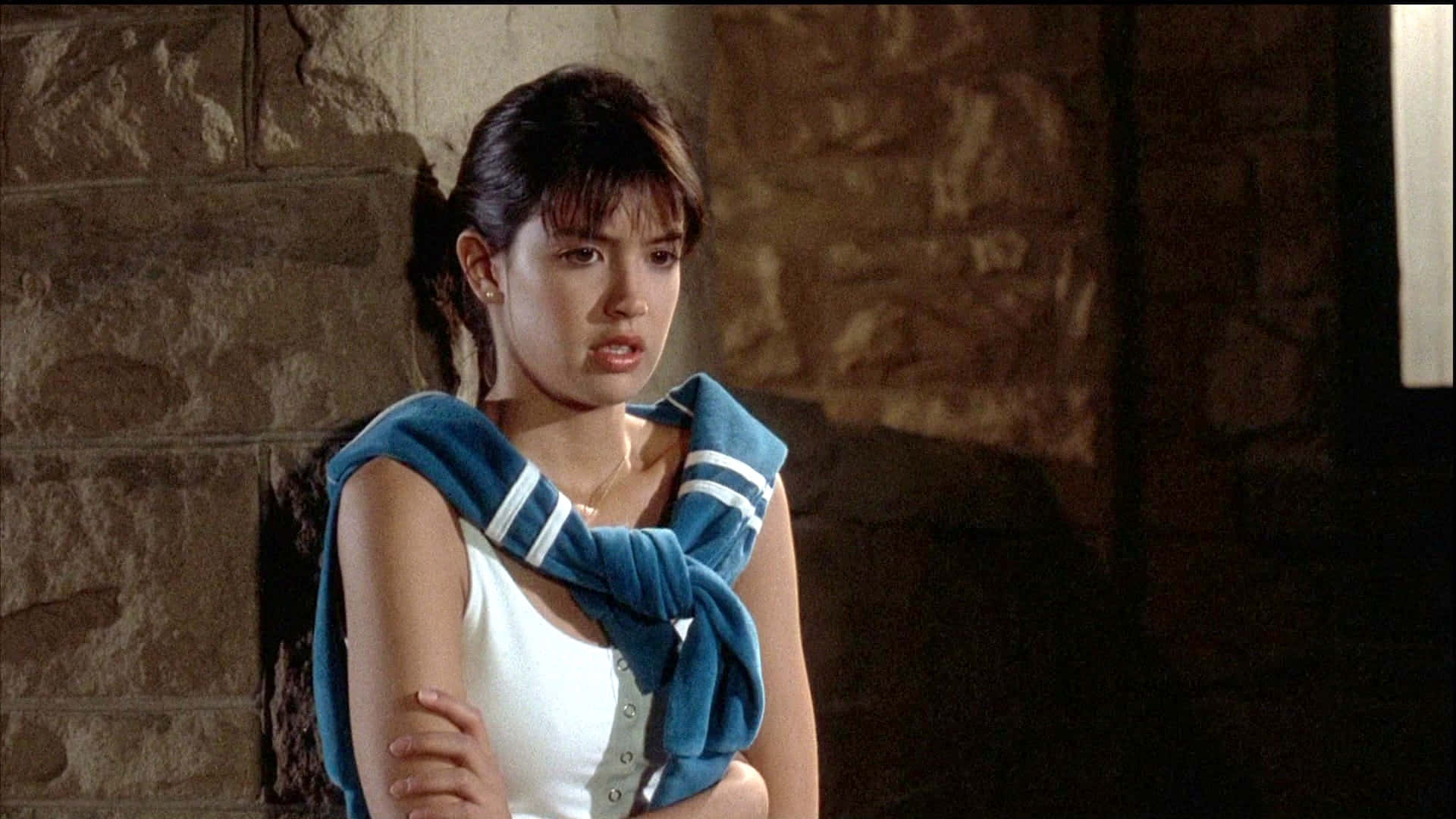 Phoebe Cates 桌布 壁紙 — 一位黑髮年輕女子靠著石牆站立，身穿白色上衣，肩上披著藍色毛巾。