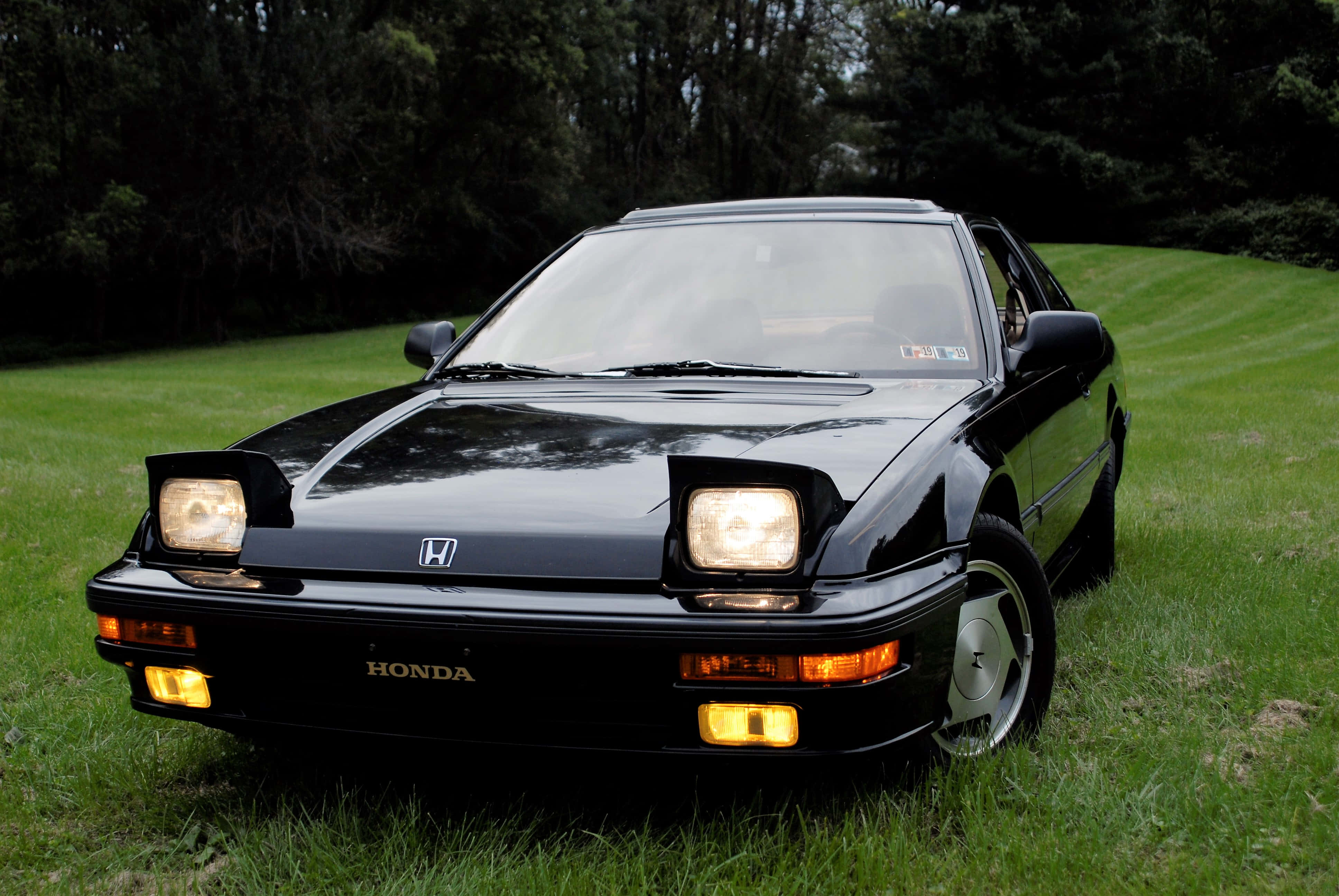 Honda Prelude 桌布 壁紙 — 一輛時尚的黑色本田跑車，車頭燈閃閃發光，停放在茂密樹木環繞的翠綠草坪上。
