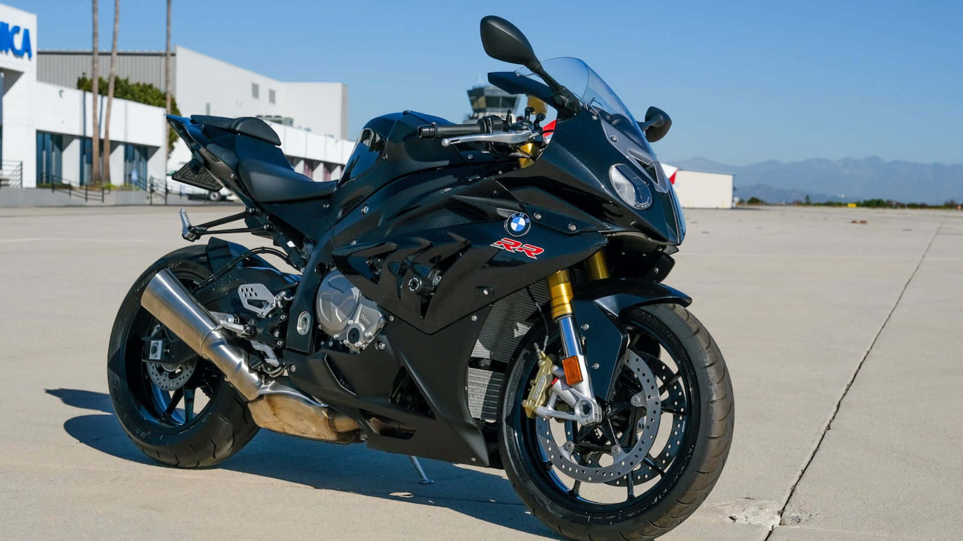  壁紙 — 一輛流線型黑色BMW R1200RR運動機車在明亮陽光下閃耀，停在混凝土停機坪上，背景有遠山和管制塔。