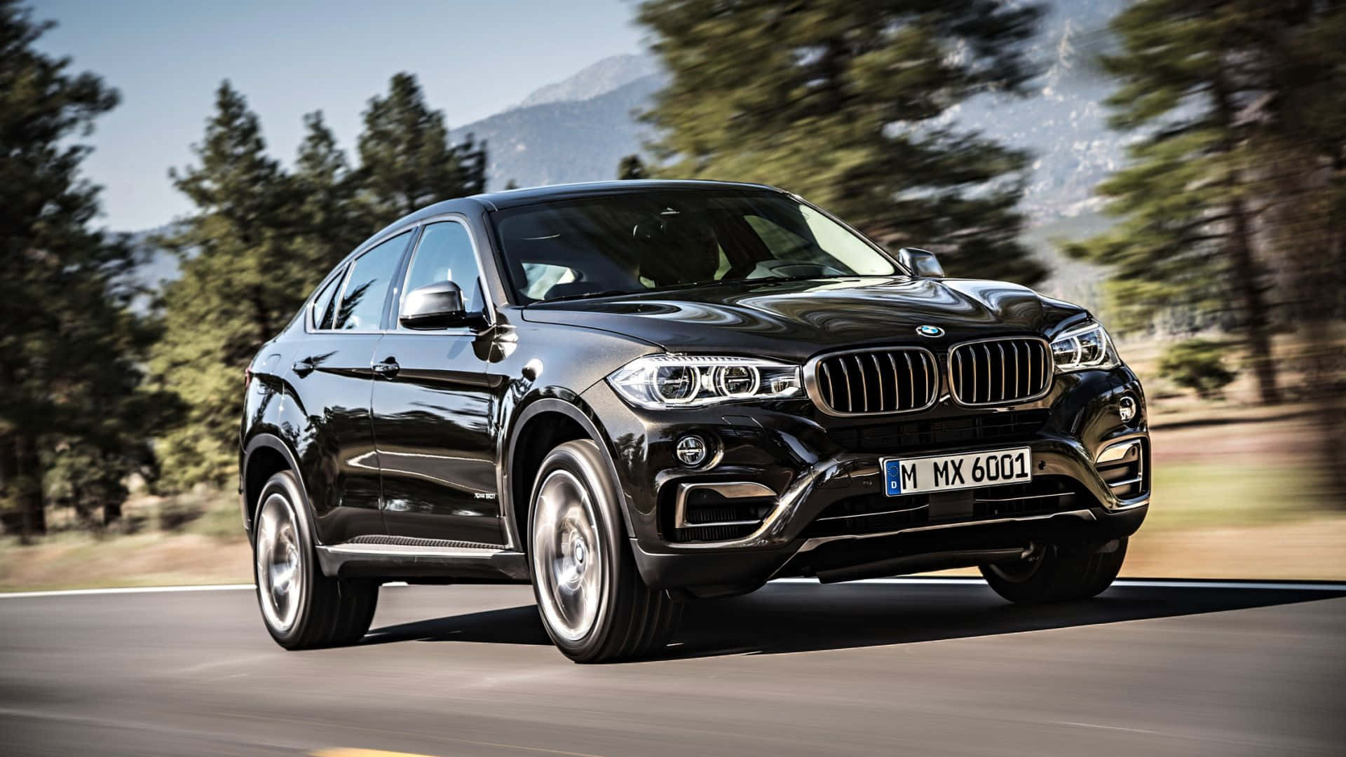 Bmw X6 桌布 壁紙 — 一輛時尚的黑色BMW X6 SUV在明亮的日光下，沿著兩旁茂密常綠樹木的蜿蜒山路飛馳。