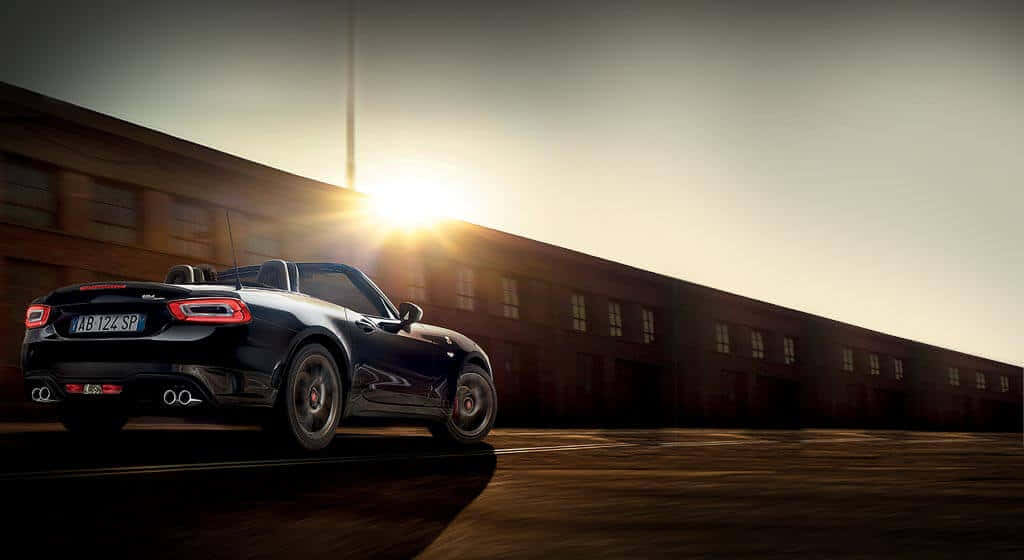 Fiat 124 Spider 桌布 壁紙 — 一輛時尚的黑色敞篷跑車在日落時停在一棟帶有發光窗戶的長工業建築旁。