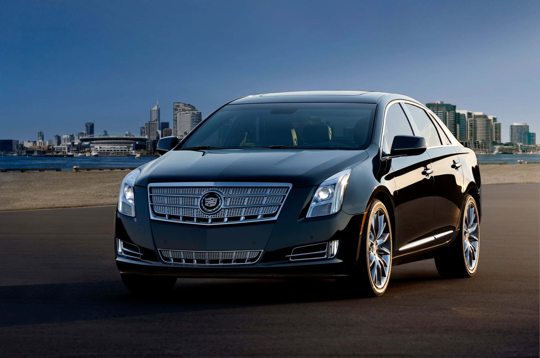 Cadillac Xts 桌布 壁紙 — 一輛時尚的黑色凱迪拉克轎車在明亮的日光下閃閃發光，停泊在海濱附近，背景是現代化的城市天際線。