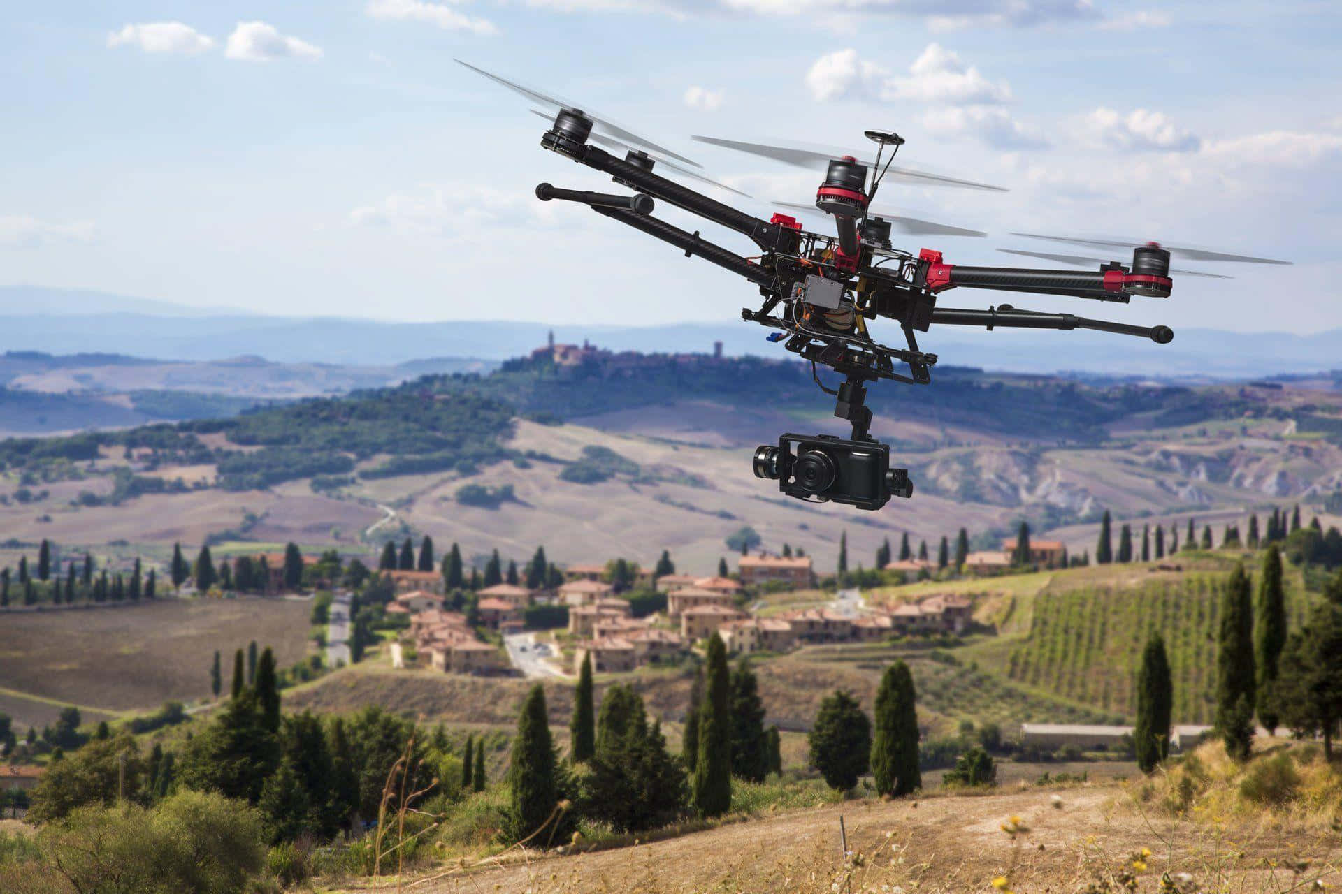 Drone Camera 桌布 壁紙 — 一架帶有紅色點綴的黑色無人機在陽光明媚的托斯卡尼鄉村上空盤旋，下方是起伏的丘陵、葡萄園和遠處山頂上的村莊，天空湛藍，白雲點點。