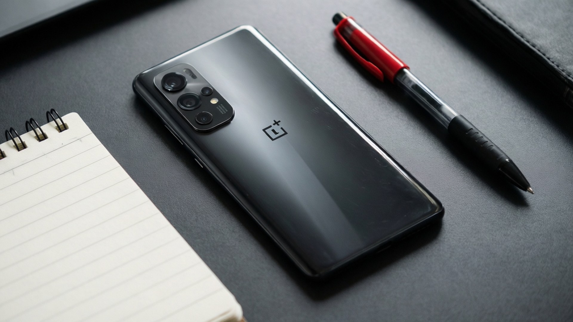 Workspace Minimal 壁紙 — 一部時尚的黑色OnePlus智慧型手機放在深灰色桌面上，旁邊有一支紅色筆和螺旋筆記本。