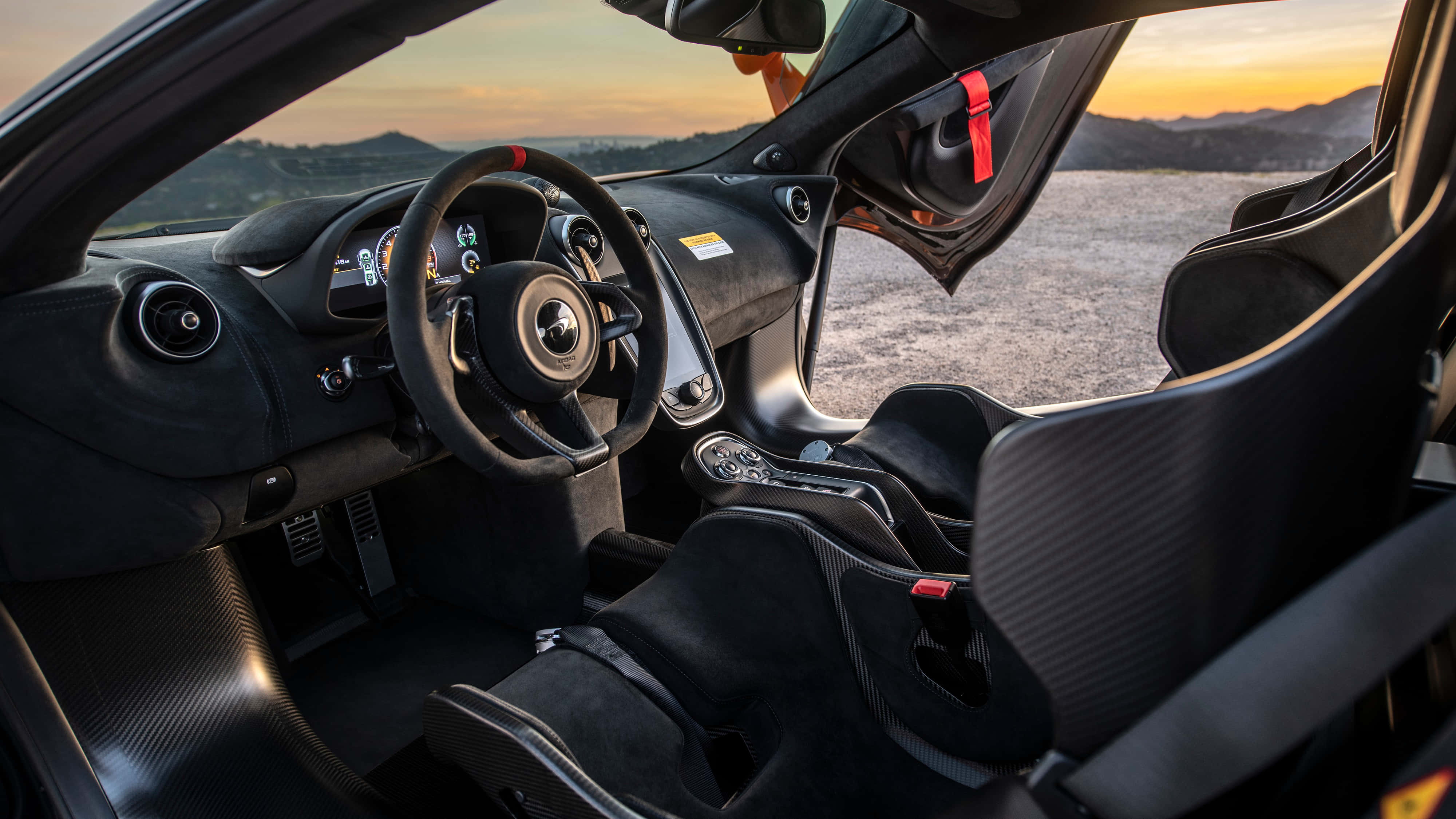 Mclaren 620r 桌布 壁紙 — 黑色跑車內飾，帶有碳纖維細節和紅色點綴，日落時分俯瞰沙漠景觀。