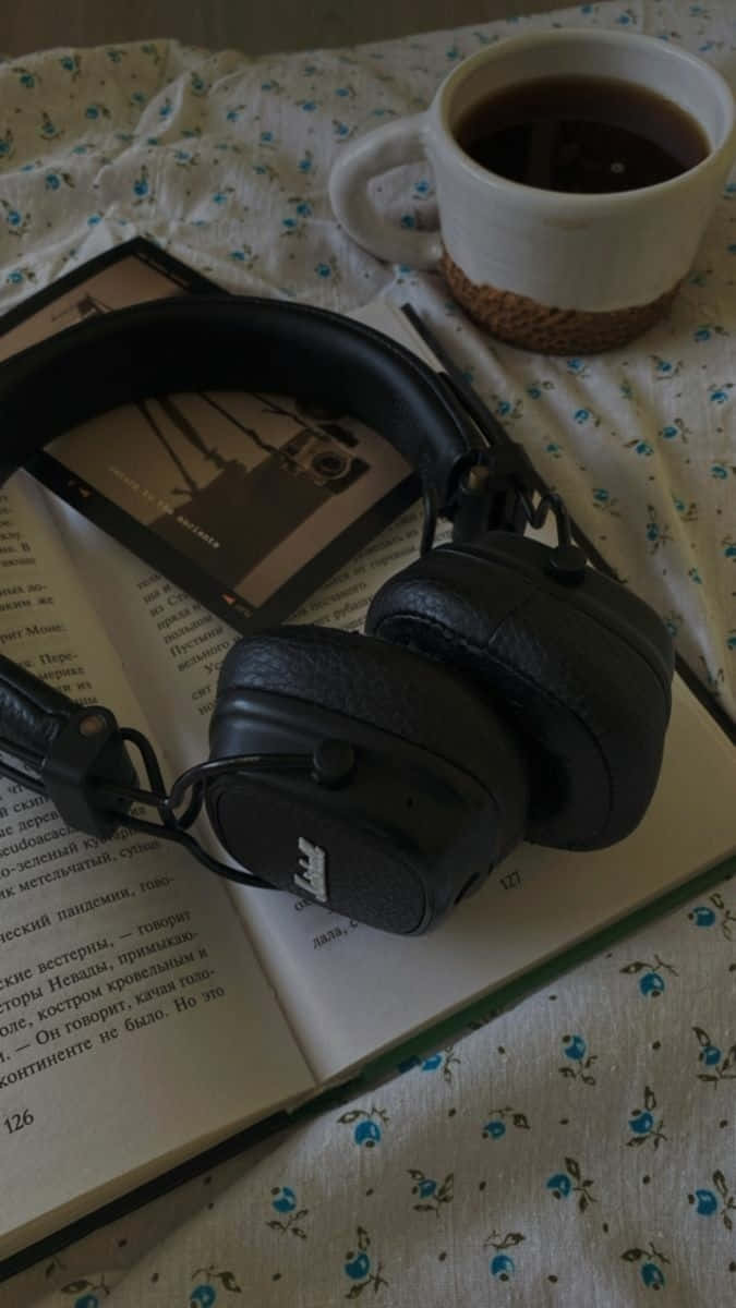  壁紙 — 黑色頭戴式耳機放在一本打開的書上，旁邊有一杯白色咖啡杯，放在花卉圖案的床罩上。