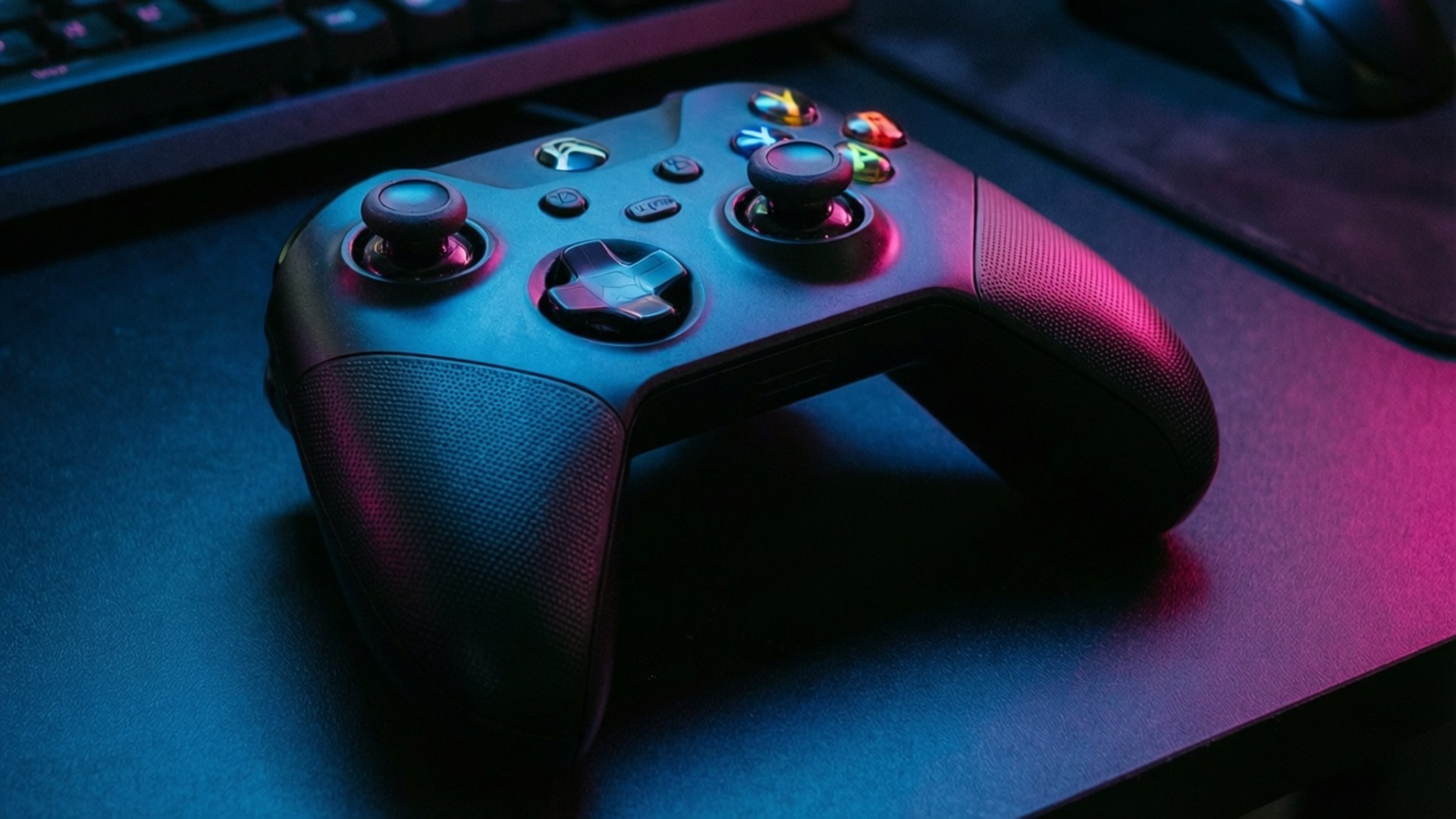 RGB Gaming Controller 壁紙 — 一個黑色Xbox手把沐浴在鮮豔的藍色和粉紅色霓虹燈光中，放在一個黑暗的桌子上，旁邊有鍵盤和滑鼠。