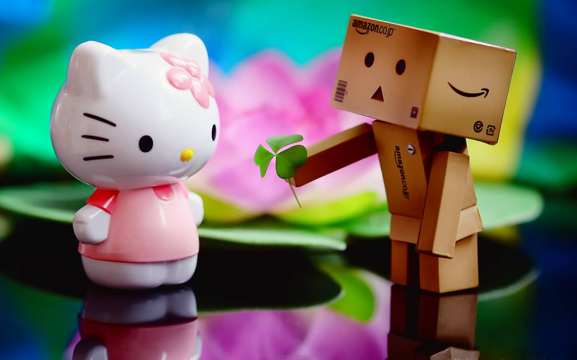 Hello 桌布 壁紙 — Hello Kitty 公仔在色彩繽紛模糊的花卉背景前，從一個微笑的紙板 Danbo 機器人那裡收下四葉草。