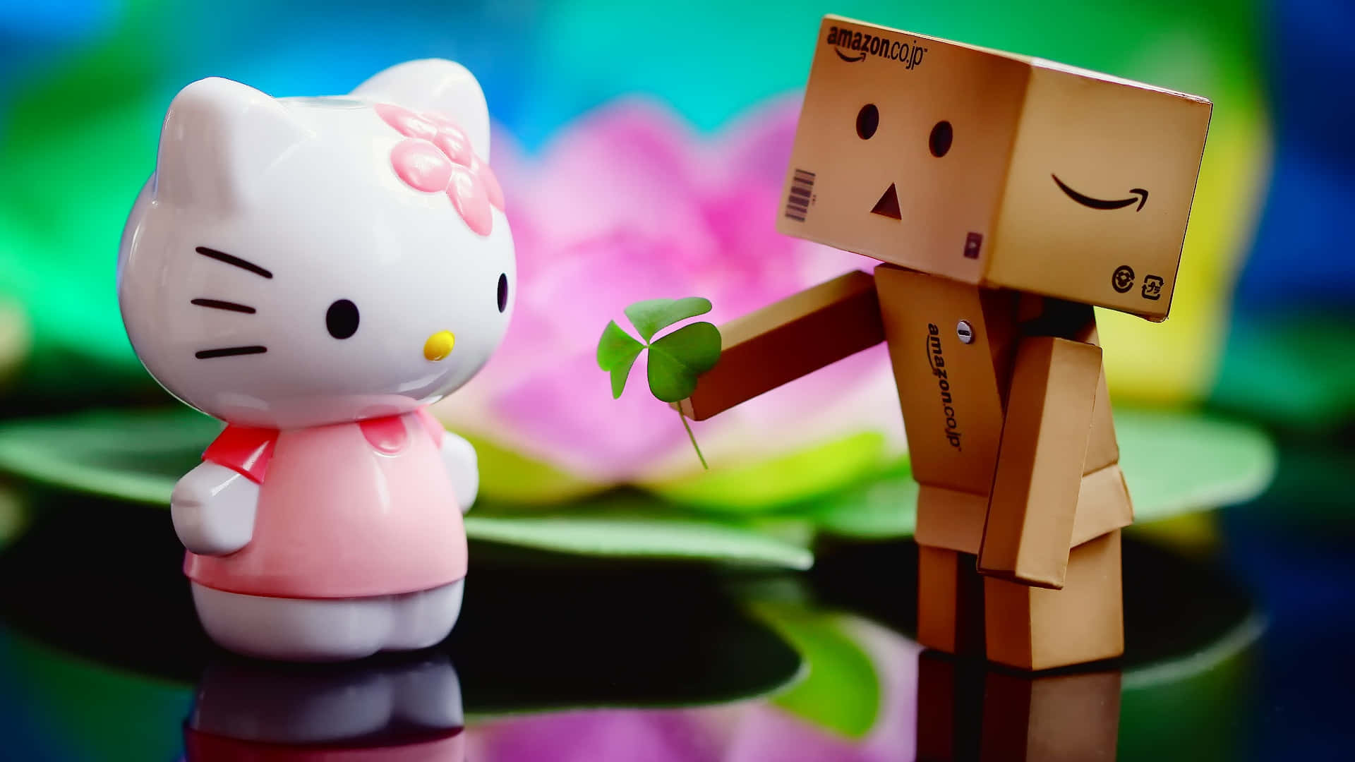 Cute Love 桌布 壁紙 — Hello Kitty 公仔從一個微笑的紙板亞馬遜盒子機器人手中接過四葉草，背景是色彩繽紛、模糊的花卉圖案。