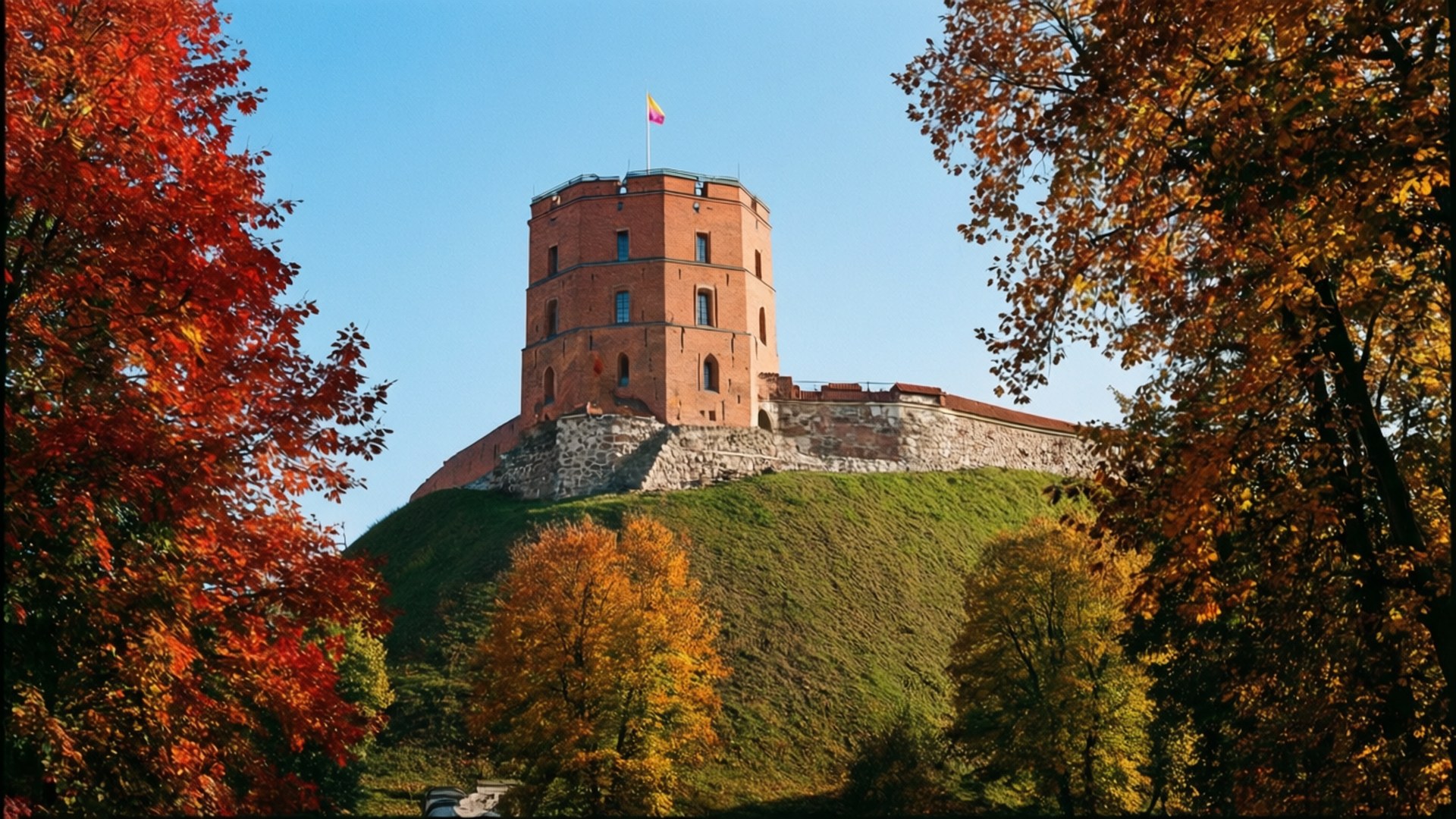 Autumn at Gediminas Tower 壁紙 — 一座歷史悠久的紅磚城堡塔樓矗立在青草覆蓋的山丘上，周圍環繞著色彩繽紛的秋樹，背景是清澈的藍天。