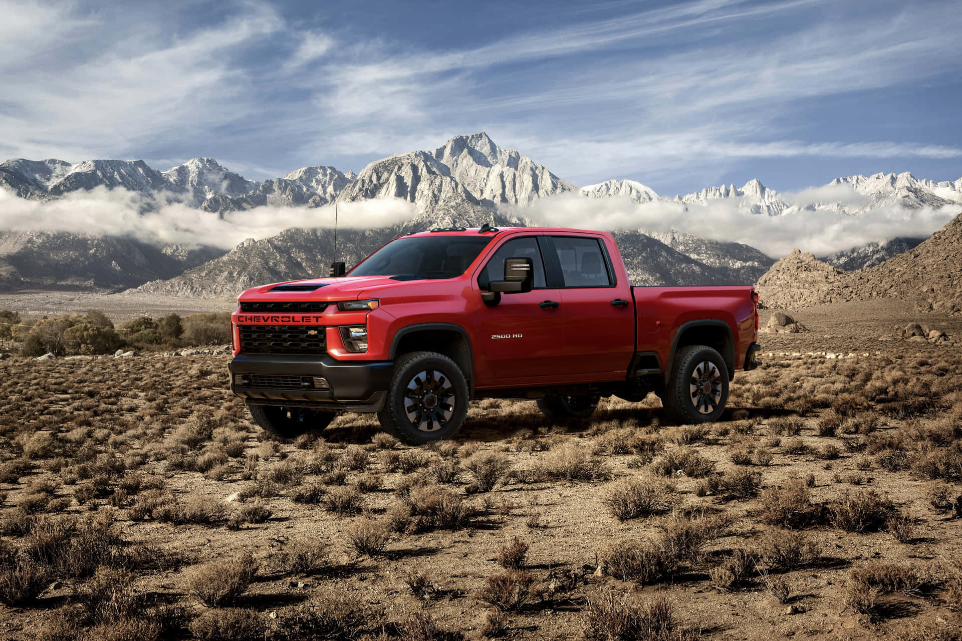 壁紙 — 一架大膽的紅色 Chevrolet Silverado 貨車停在乾燥的沙漠田野中，背後是部分多雲的天空下積雪覆蓋的山脈。