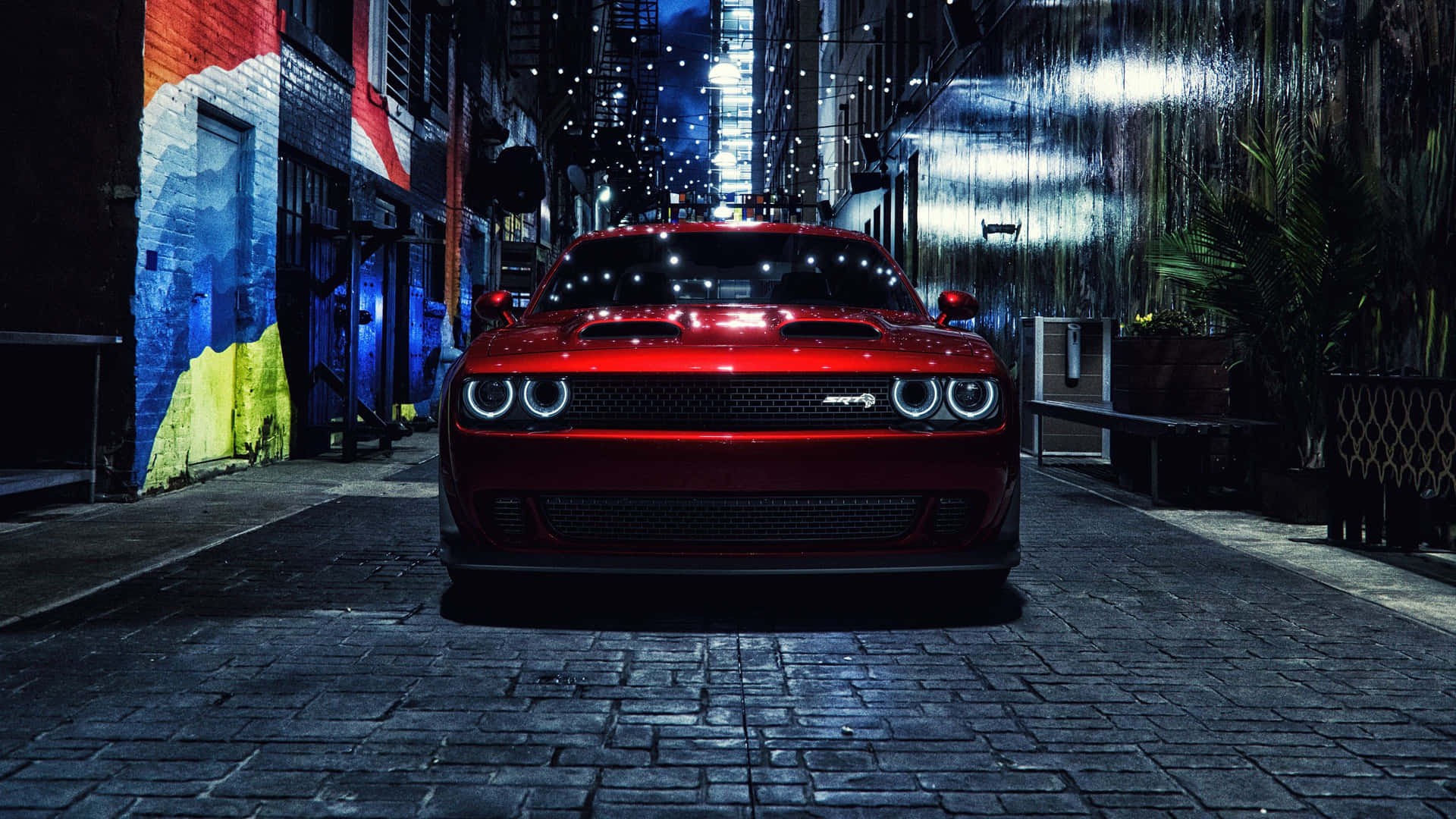  壁紙 — 一輛閃亮的紅色 Dodge Challenger SRT Hellcat 在夜間主宰著一條狹窄的鵝卵石小巷，兩旁是色彩繽紛的塗鴉和發光的燈串。