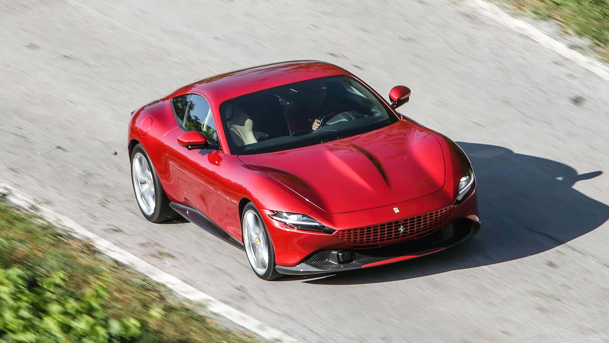 Ferrari Roma 桌布 壁紙 — 一輛鮮紅色的法拉利跑車在明亮的陽光下，沿著蜿蜒的道路疾馳，周圍是模糊的綠化和瀝青。