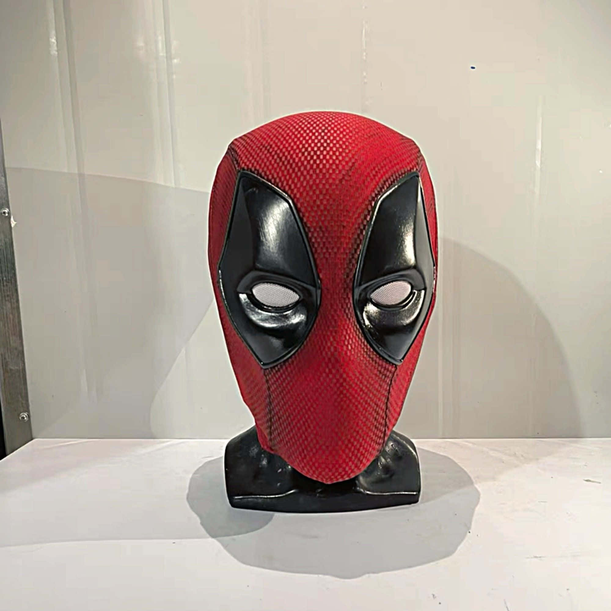 Deadpool Mask 桌布 壁紙 — 一個紅色和黑色的死侍面具，帶有紋理布料和光澤眼縫，放在黑色支架上，背景是純白牆壁。