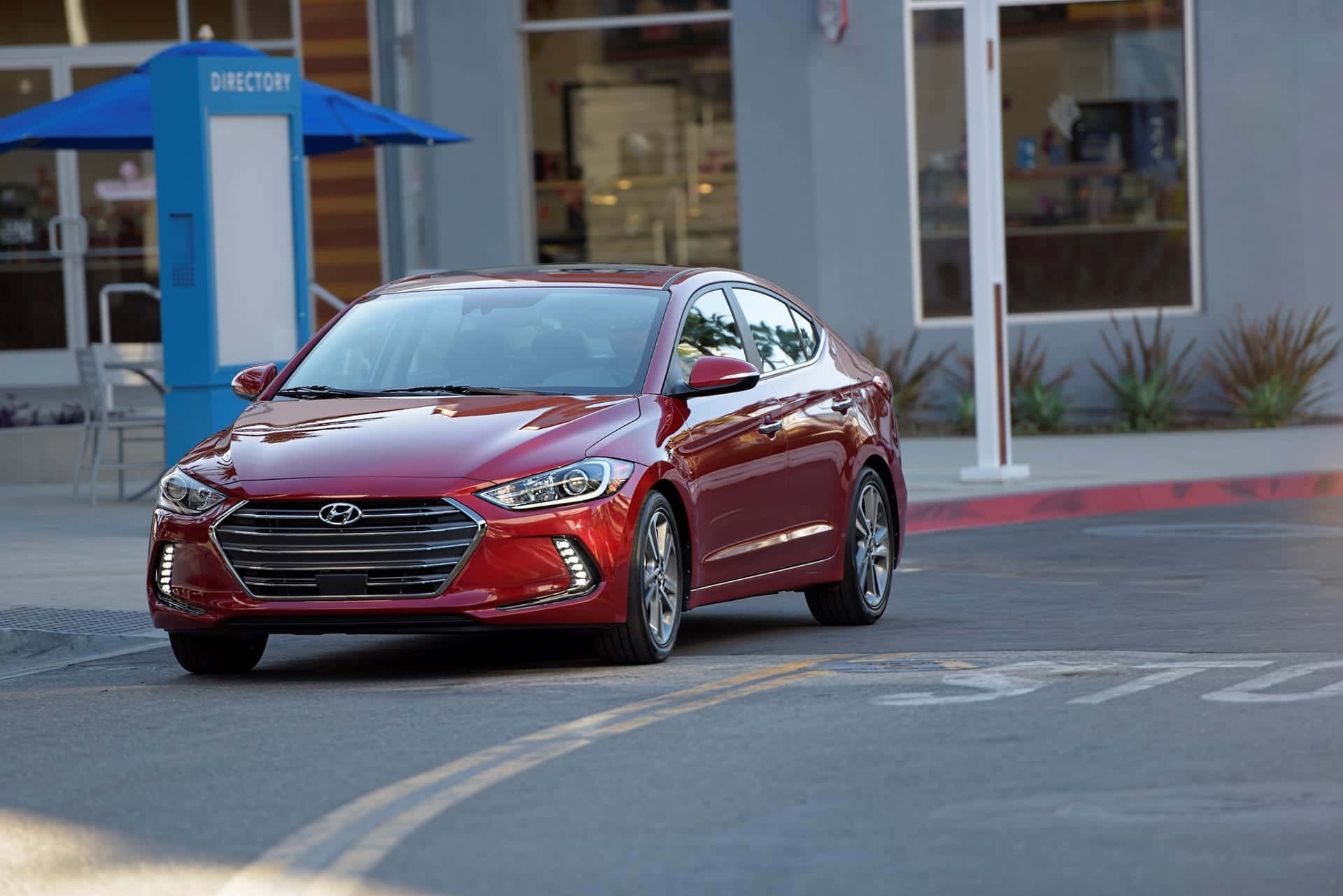 Hyundai Elantra 桌布 壁紙 — 一輛光亮的紅色現代轎車停放在加油站，背景有藍色目錄亭和商店門面。
