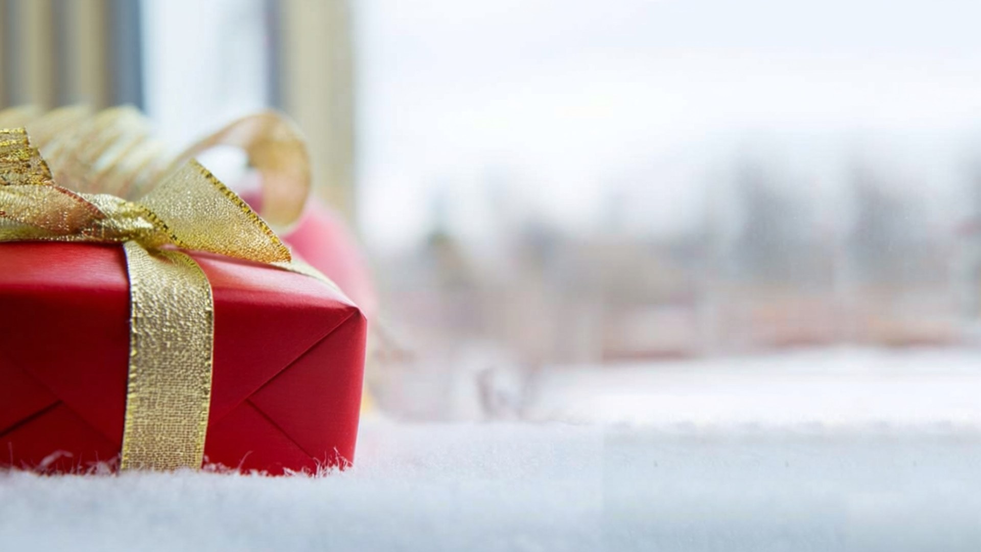 Red Ribbon Gift 壁紙 — 一個紅色禮物盒，上面有金色蝴蝶結，放在雪地上，旁邊是模糊的窗戶。