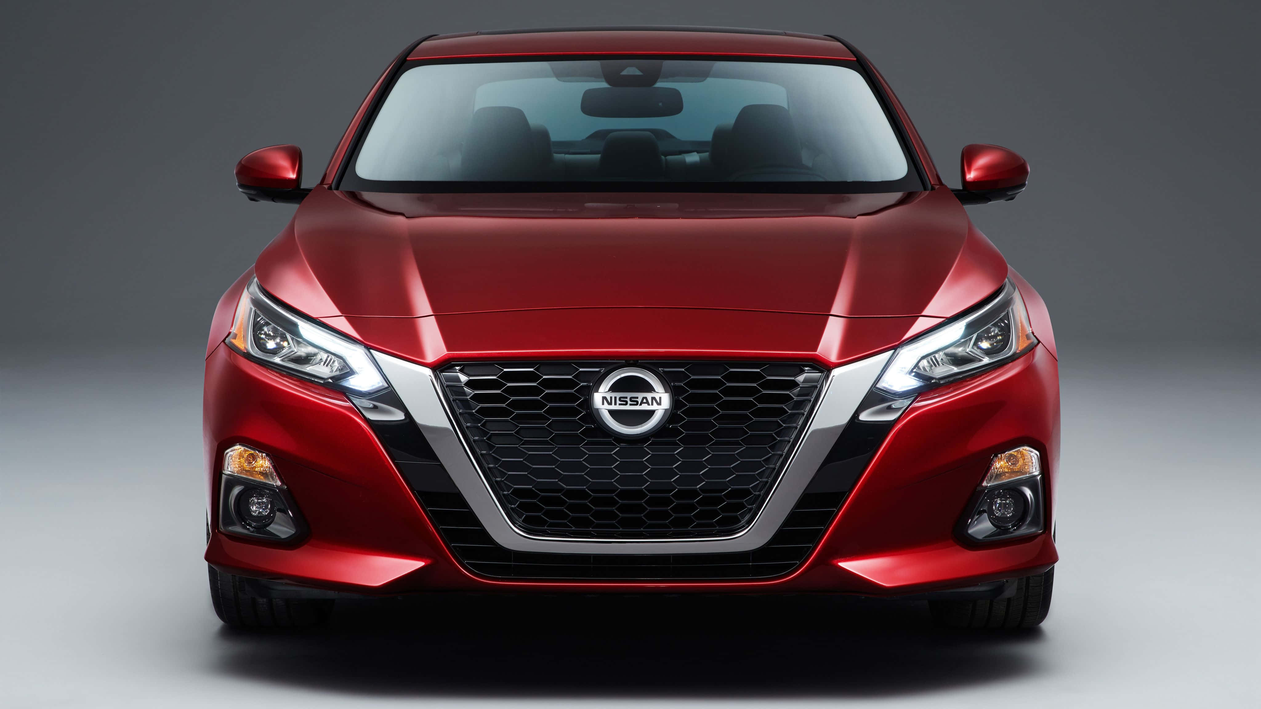 Nissan Altima 桌布 壁紙 — 一輛光亮的紅色日產轎車正面朝前，在攝影棚內以戲劇性燈光突顯其時尚現代的水箱護罩和銳利頭燈。