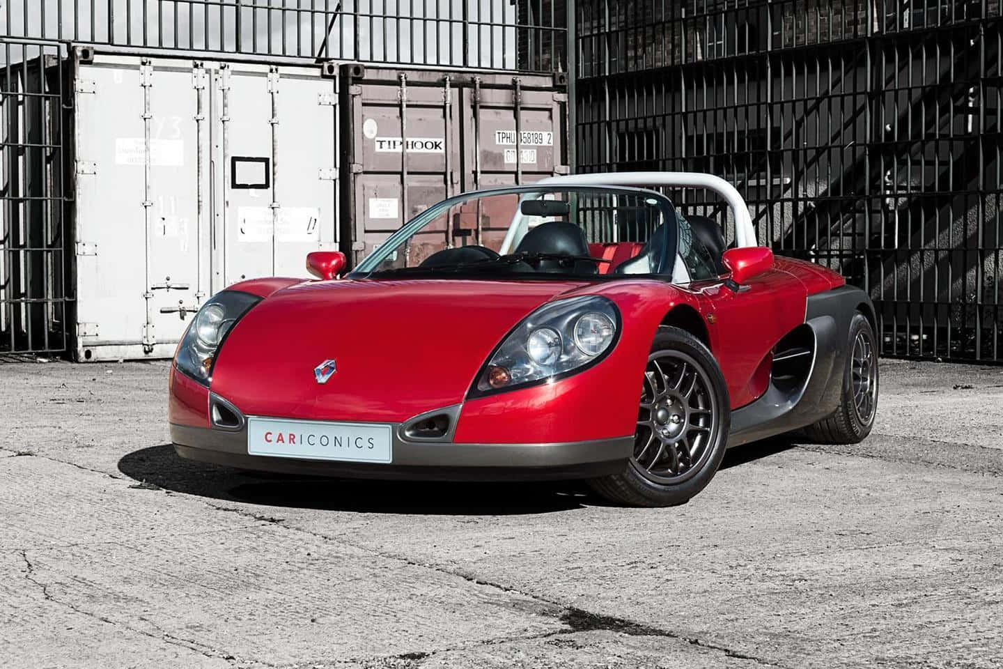 Renault Sport Spider 桌布 壁紙 — 一輛充滿活力的紅色跑車，帶有黑色飾邊和白色防滾架，停在裂開的混凝土上，前面是貨運貨櫃和黑色金屬圍欄。