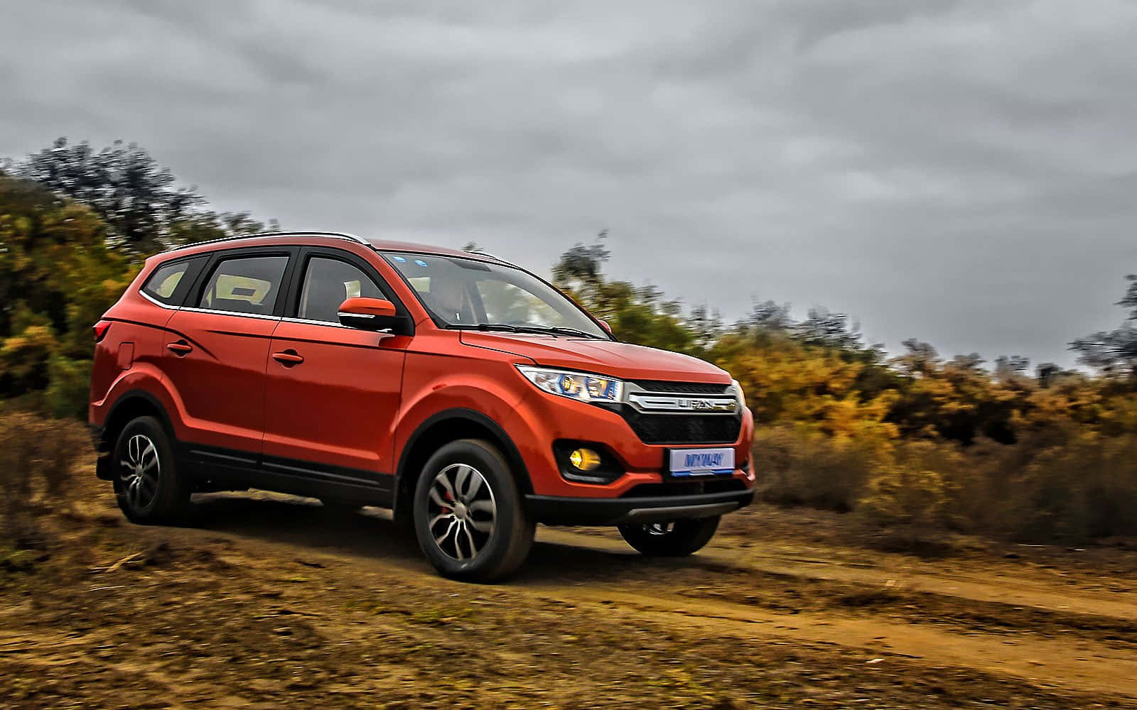  壁紙 — 一輛鮮紅色SUV在多雲天空下沿著泥路穿過秋葉動態行駛。
